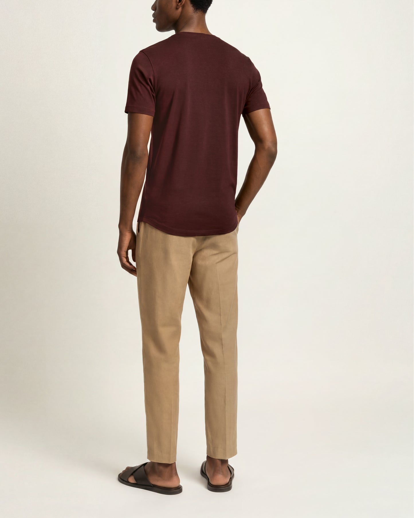 A man shown from the back wearing a short-sleeve crew-neck t-shirt in maroon, beige trousers, and dark brown slide sandals, photographed against a plain light gray studio background.