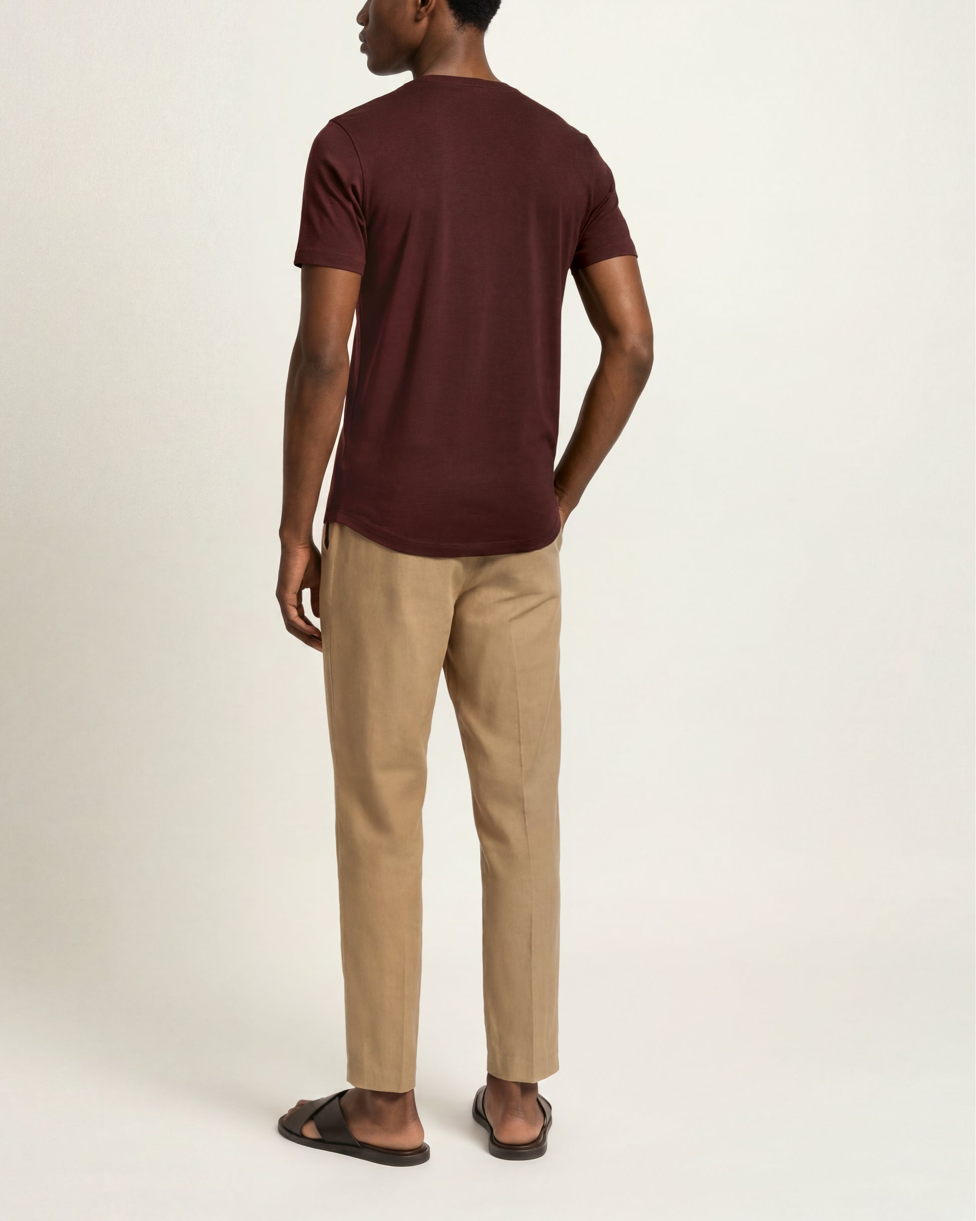 A man shown from the back wearing a short-sleeve crew-neck t-shirt in maroon, beige trousers, and dark brown slide sandals, photographed against a plain light gray studio background.