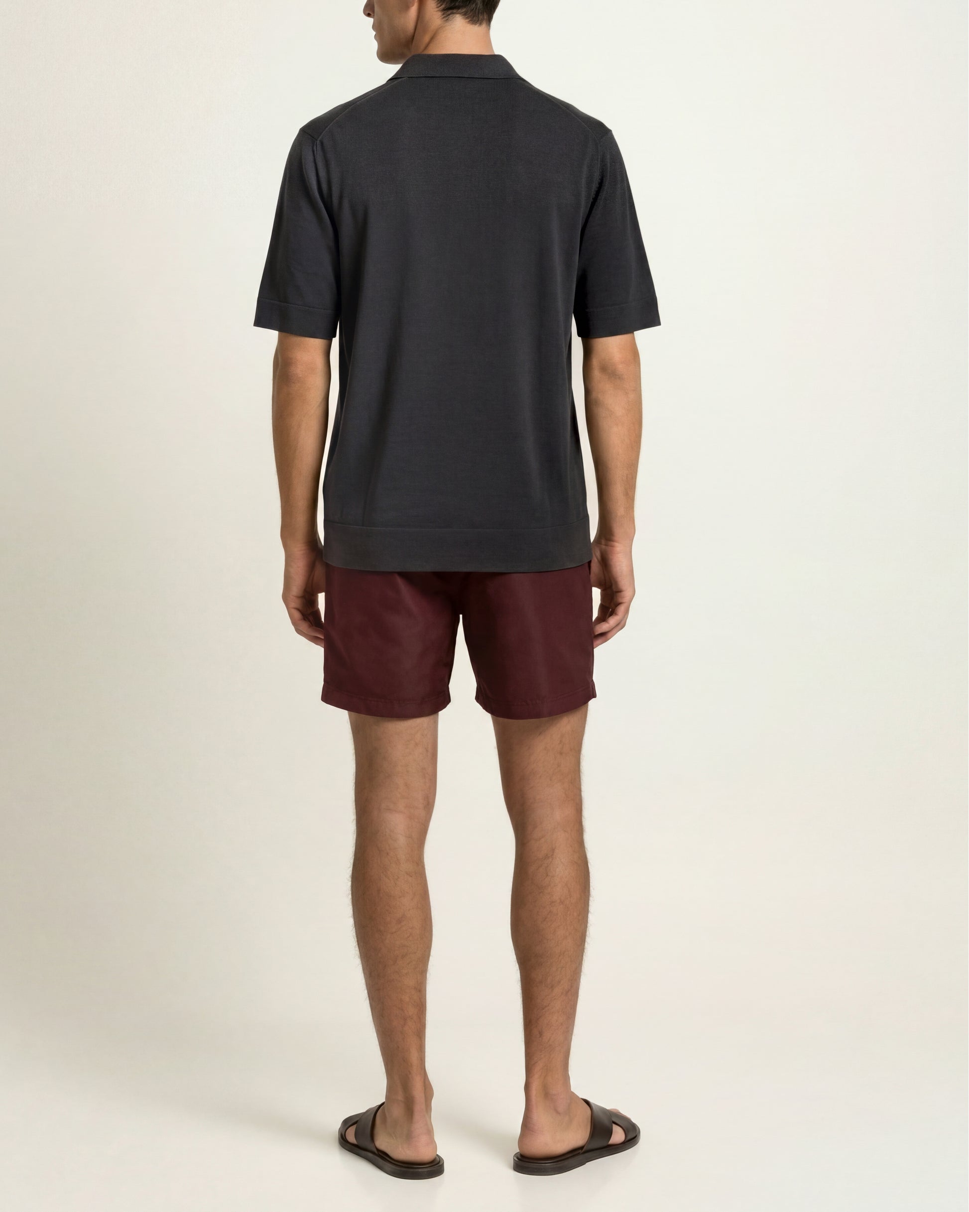 A man viewed from the back wearing a dark charcoal gray short-sleeve polo shirt and burgundy swim shorts, standing against a plain off-white studio background.