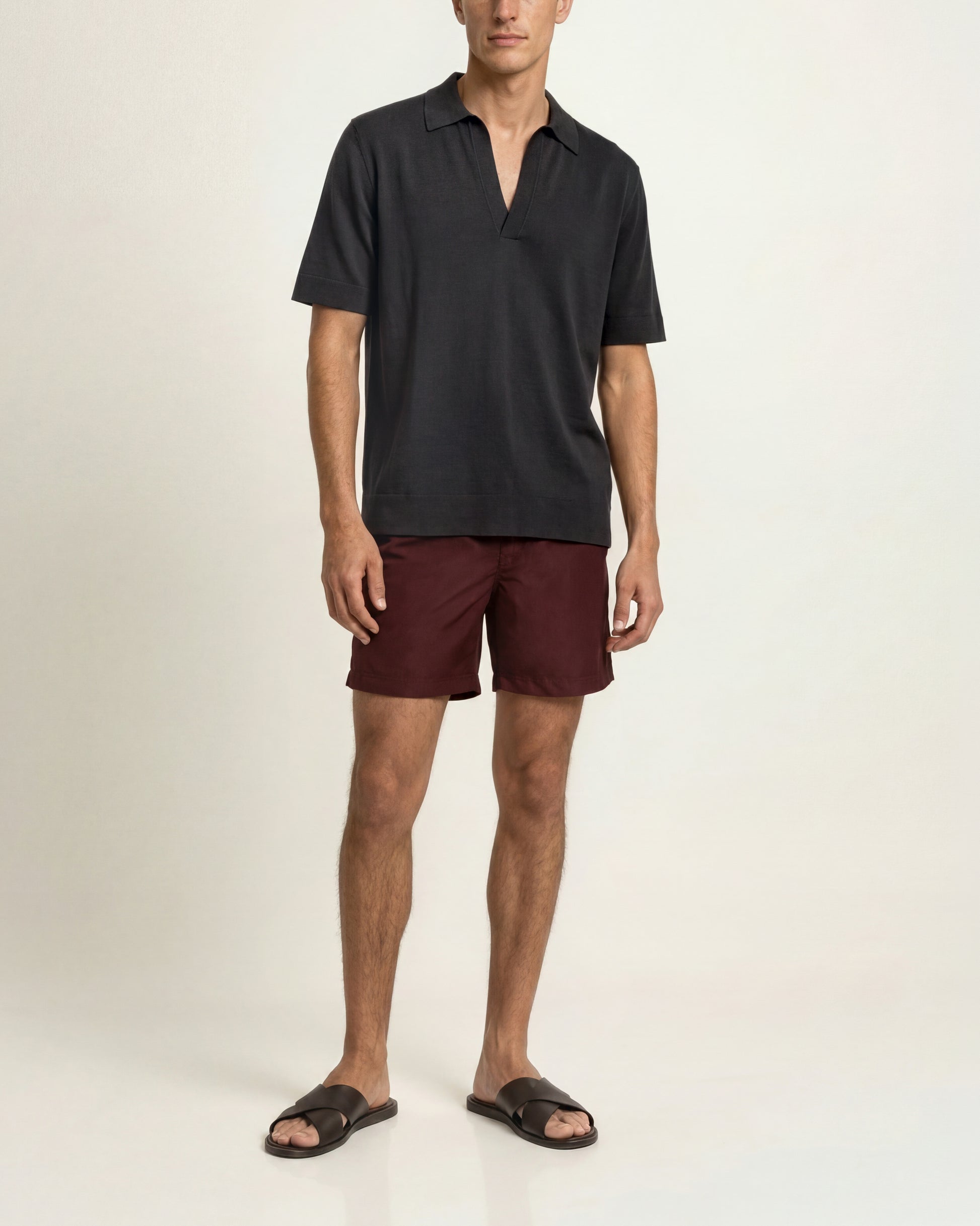 A man modeling a black short-sleeve polo shirt and burgundy shorts with black slide sandals, photographed against a plain off-white studio background.