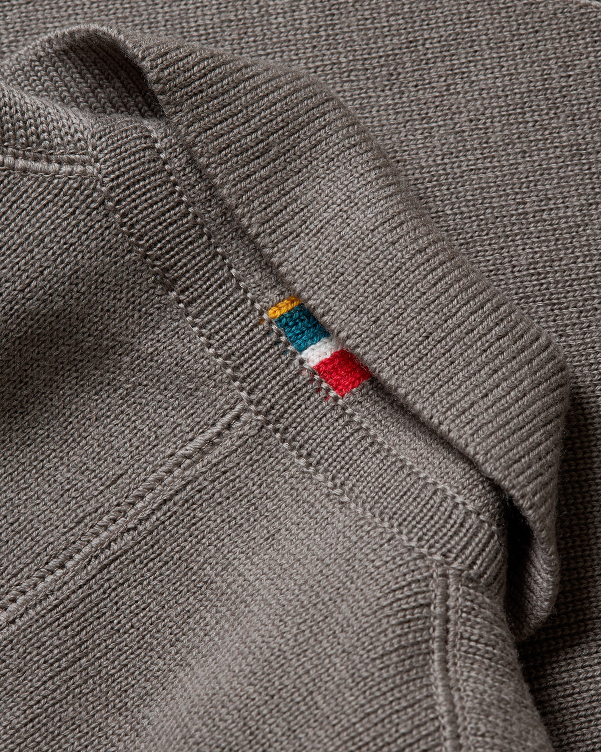 Close-up of a ribbed gray knit garment with a small colorful embroidered logo tag near the shoulder seam.