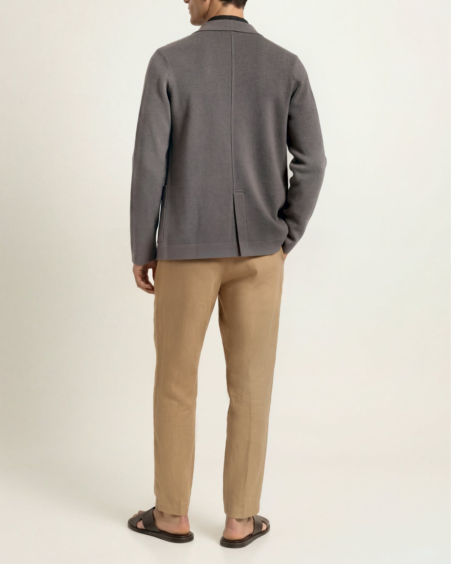 A man shown from the back wearing a dark gray knit blazer, beige trousers, and dark brown sandals, standing against a plain off-white studio background.