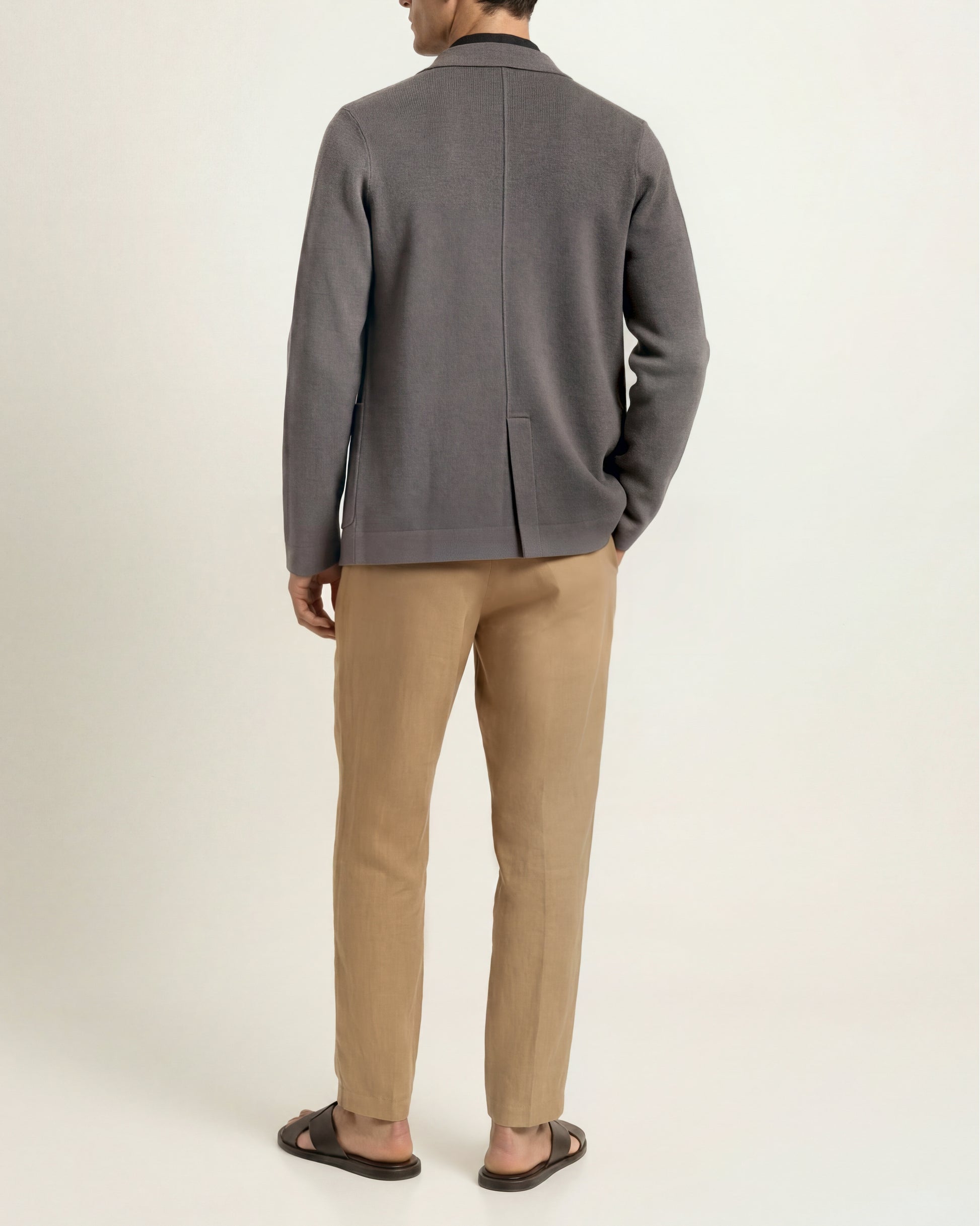 A man shown from the back wearing a dark gray knit blazer, beige trousers, and dark brown sandals, standing against a plain off-white studio background.