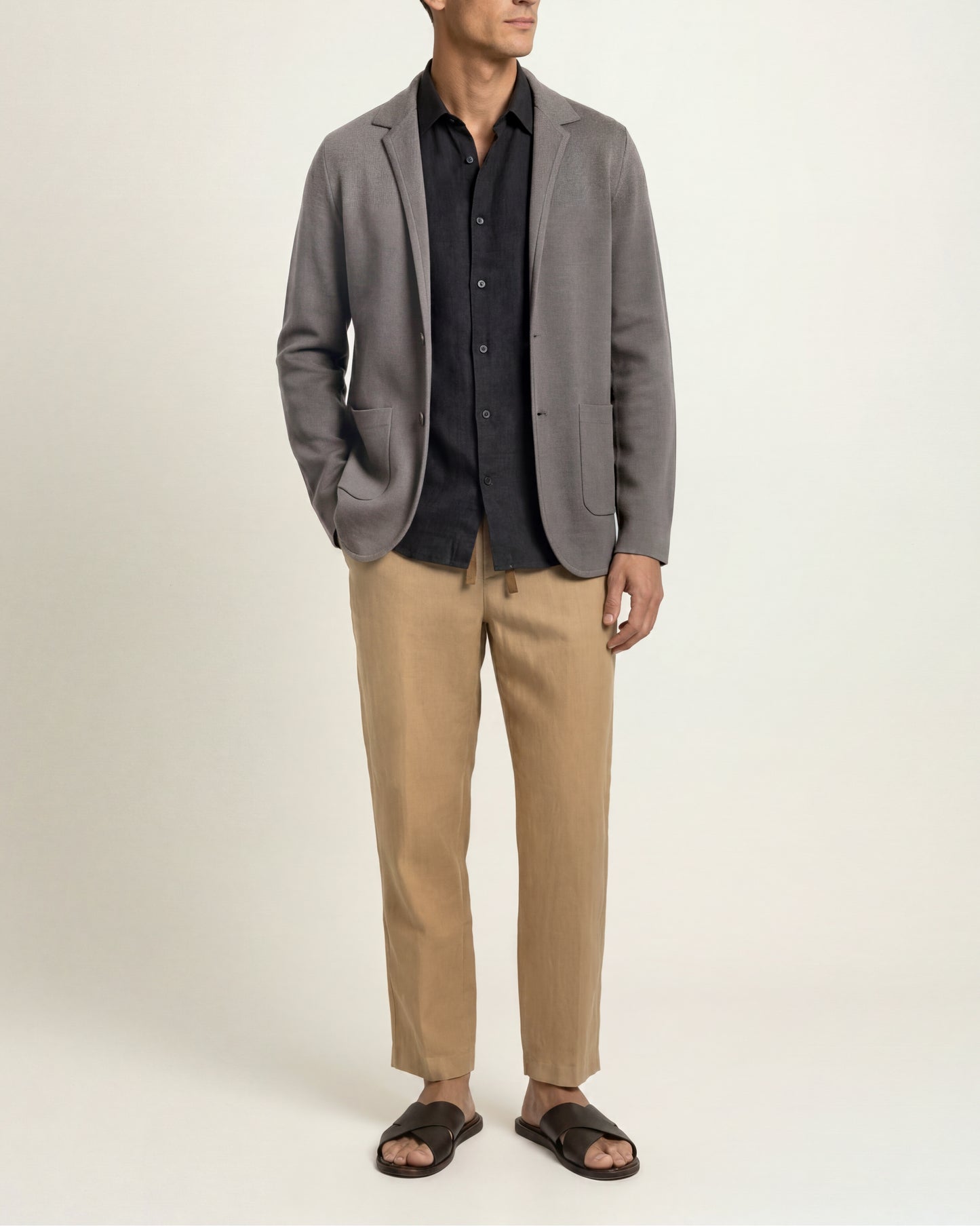 A man wearing a gray blazer over a black button-up shirt, beige trousers, and black slide sandals, standing against a plain light beige studio background.