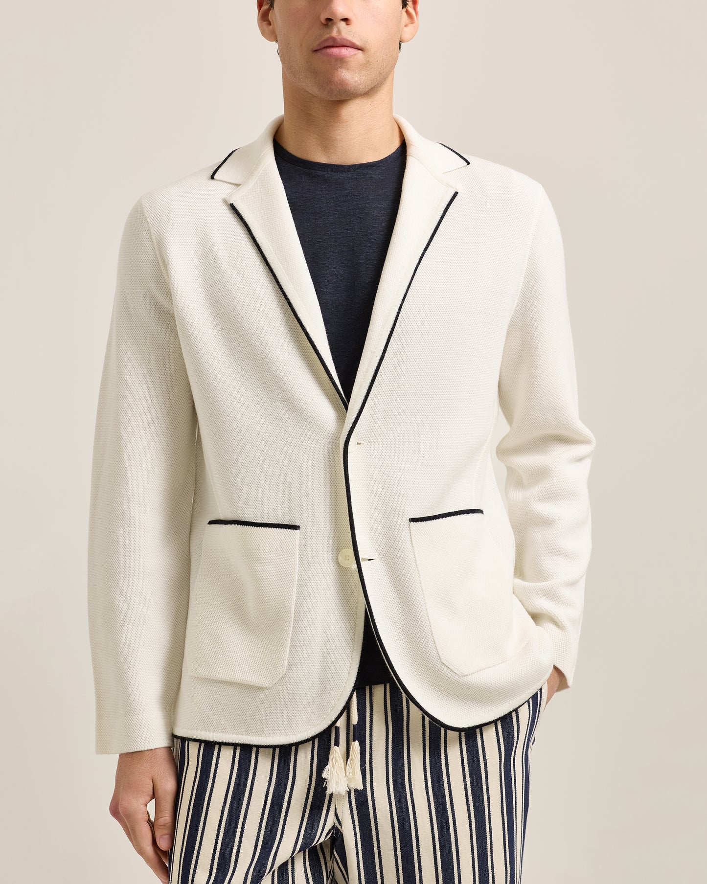 Man wearing a white blazer with black trim and striped pants on a plain background