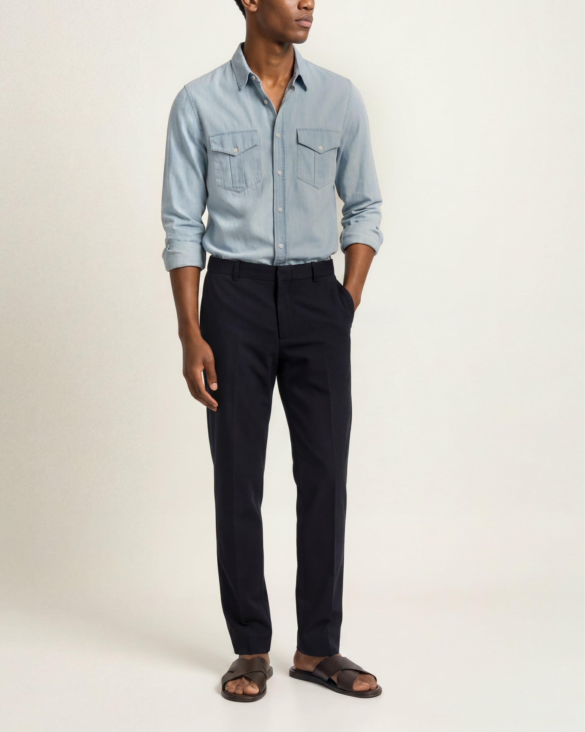 A man modeling a light blue long-sleeve button-up shirt with chest pockets, paired with dark navy trousers and brown slide sandals, against a plain off-white studio background.