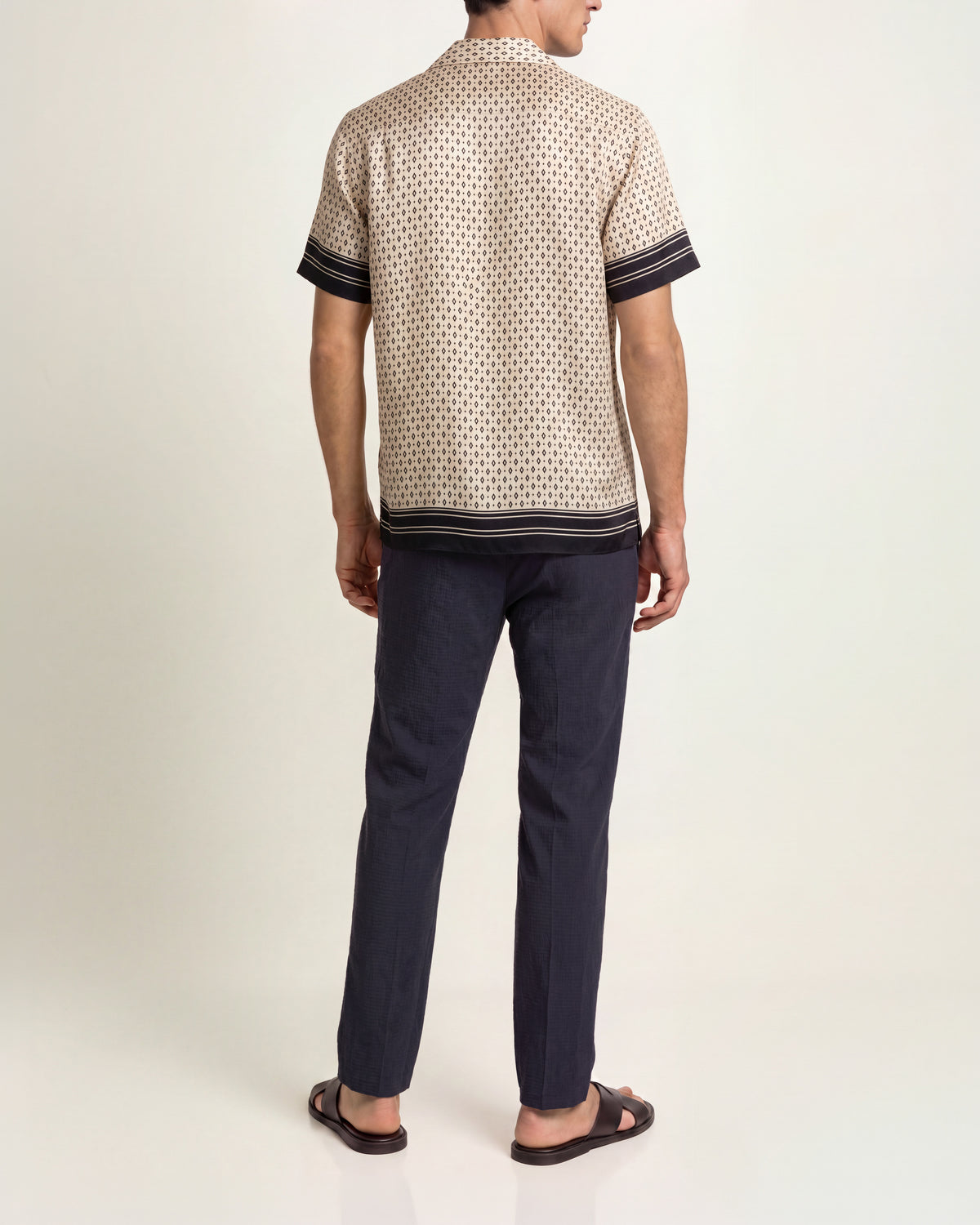 Man wearing cream and black printed silk shirt with navy trousers and brown sandals on a plain background