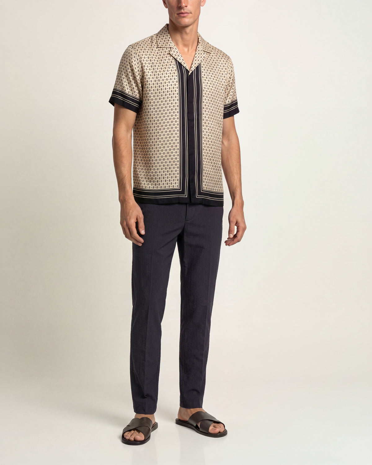 Man wearing cream and black printed silk shirt with navy trousers and brown sandals on a plain background