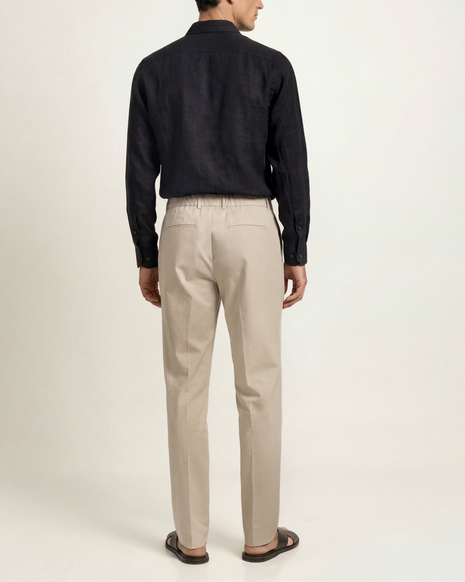 A man shown from the back wearing beige trousers, a dark navy long-sleeve shirt, and brown sandals, photographed against a plain light background.