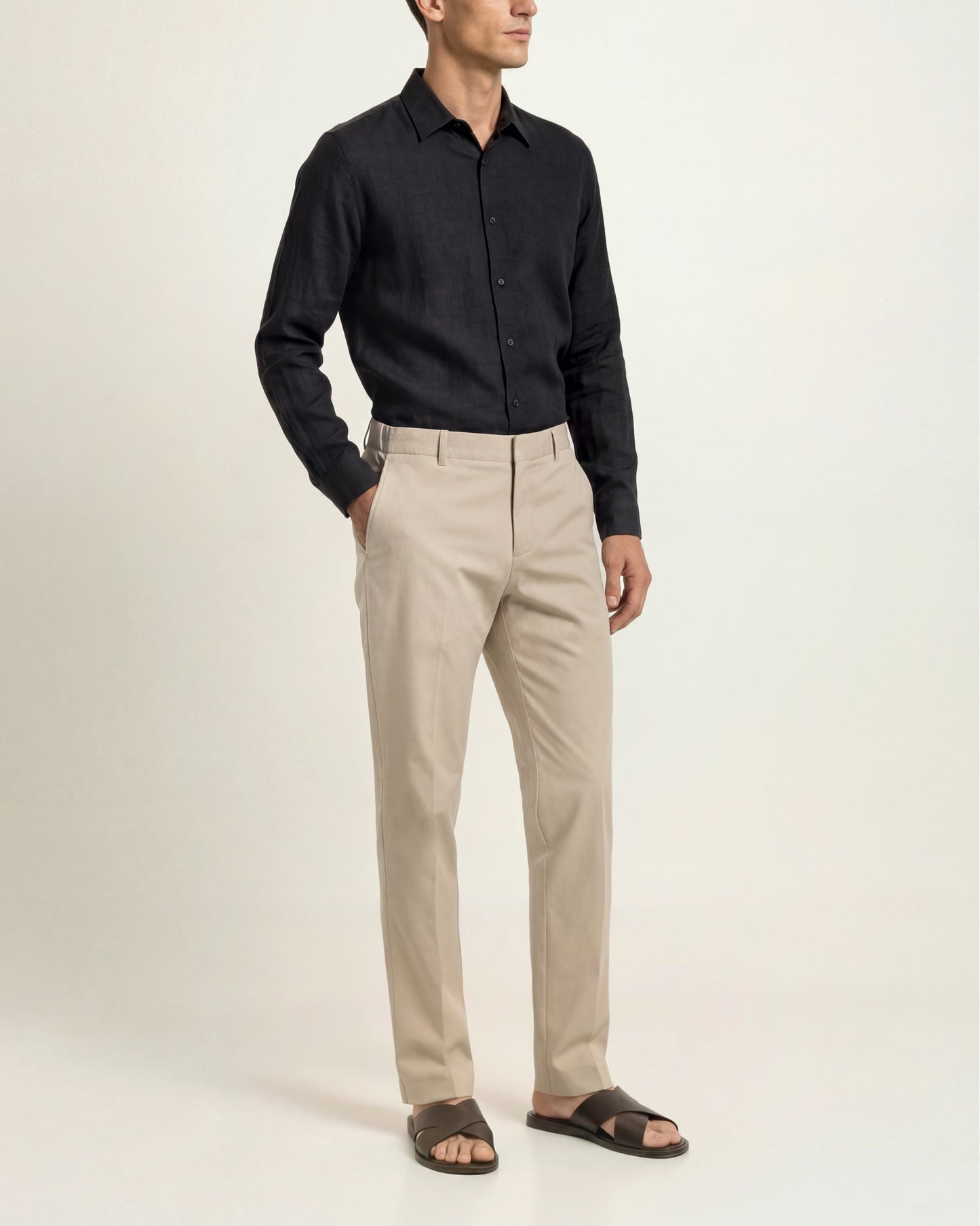 A man wearing a black long-sleeve button-down shirt, beige chinos, and dark brown slide sandals, standing against a plain off-white studio background.
