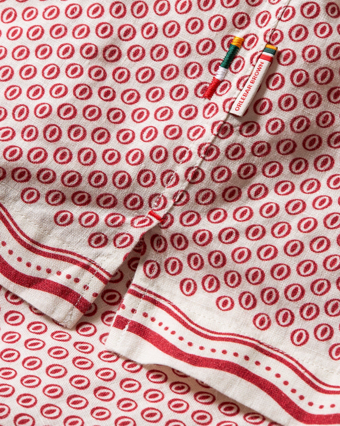 A close-up of a white cotton kitchen towel or napkin featuring a repeating red circular motif and red striped borders, with a small colorful sewing tool placed on top.