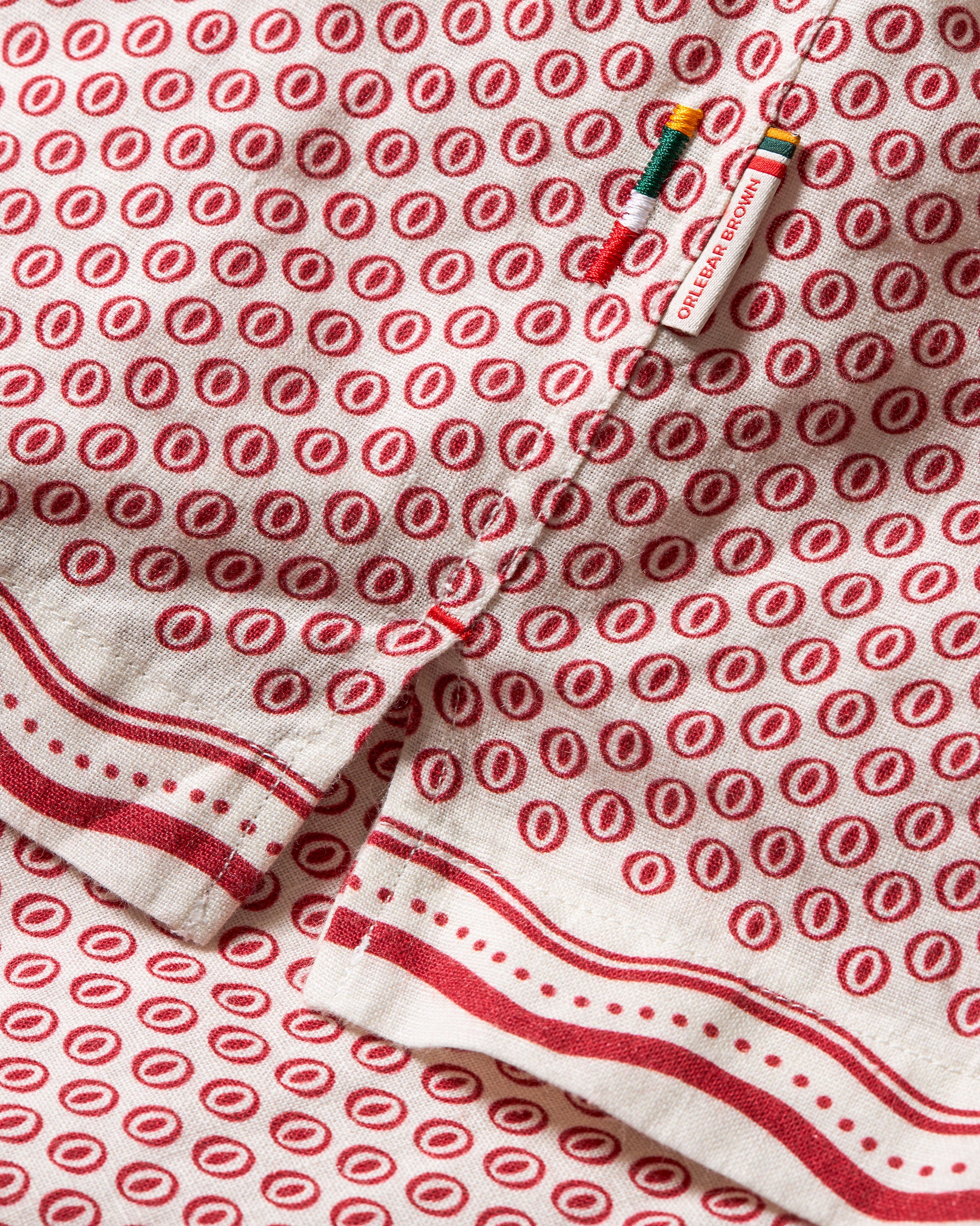 A close-up of a white cotton kitchen towel or napkin featuring a repeating red circular motif and red striped borders, with a small colorful sewing tool placed on top.