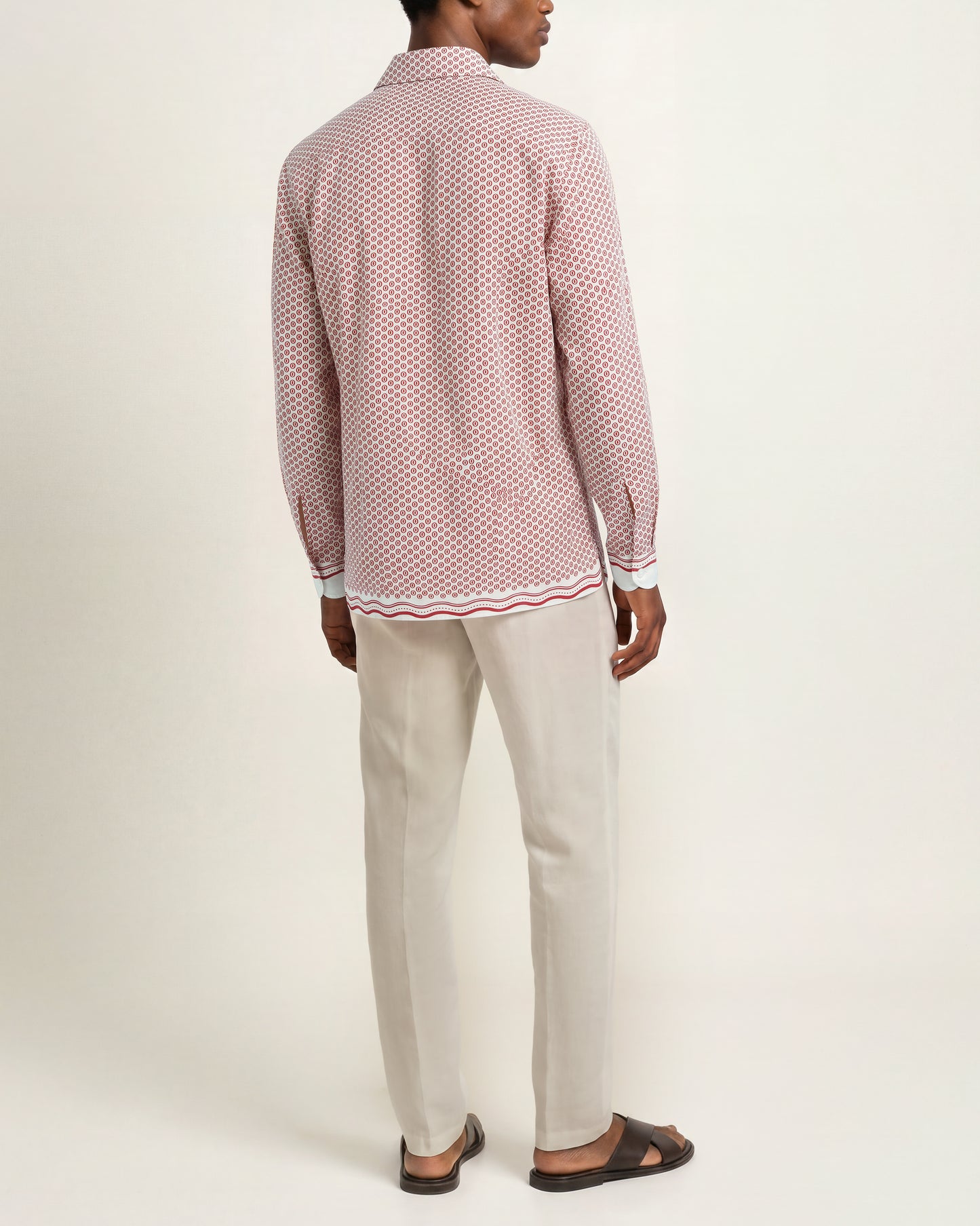 A man shown from the back wearing a pink and white patterned long-sleeve shirt, beige trousers, and dark brown sandals against a plain light background.