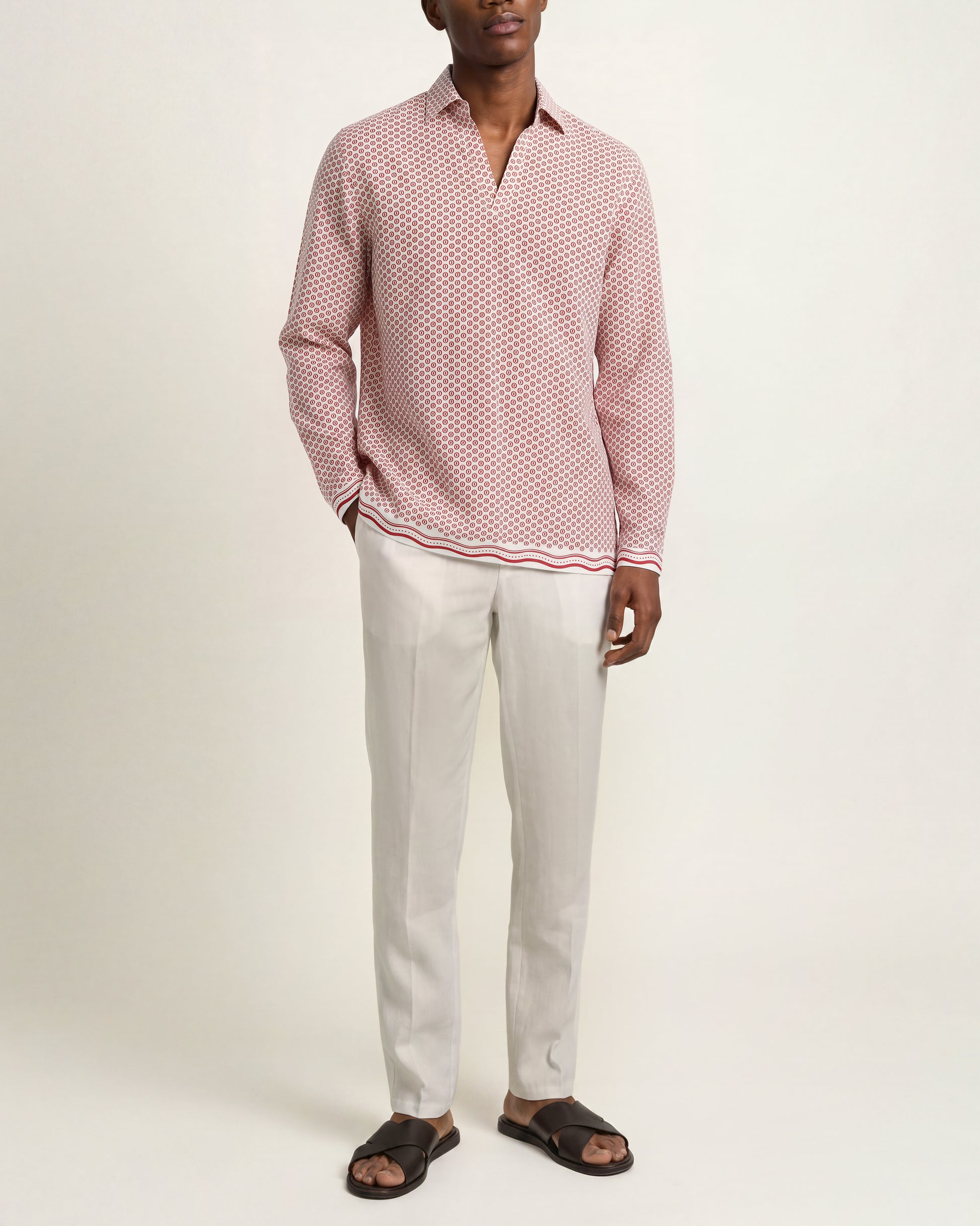 A man modeling a pink and white micro-patterned long-sleeve polo shirt with a striped hem, paired with off-white trousers and black slide sandals, against a plain light background.
