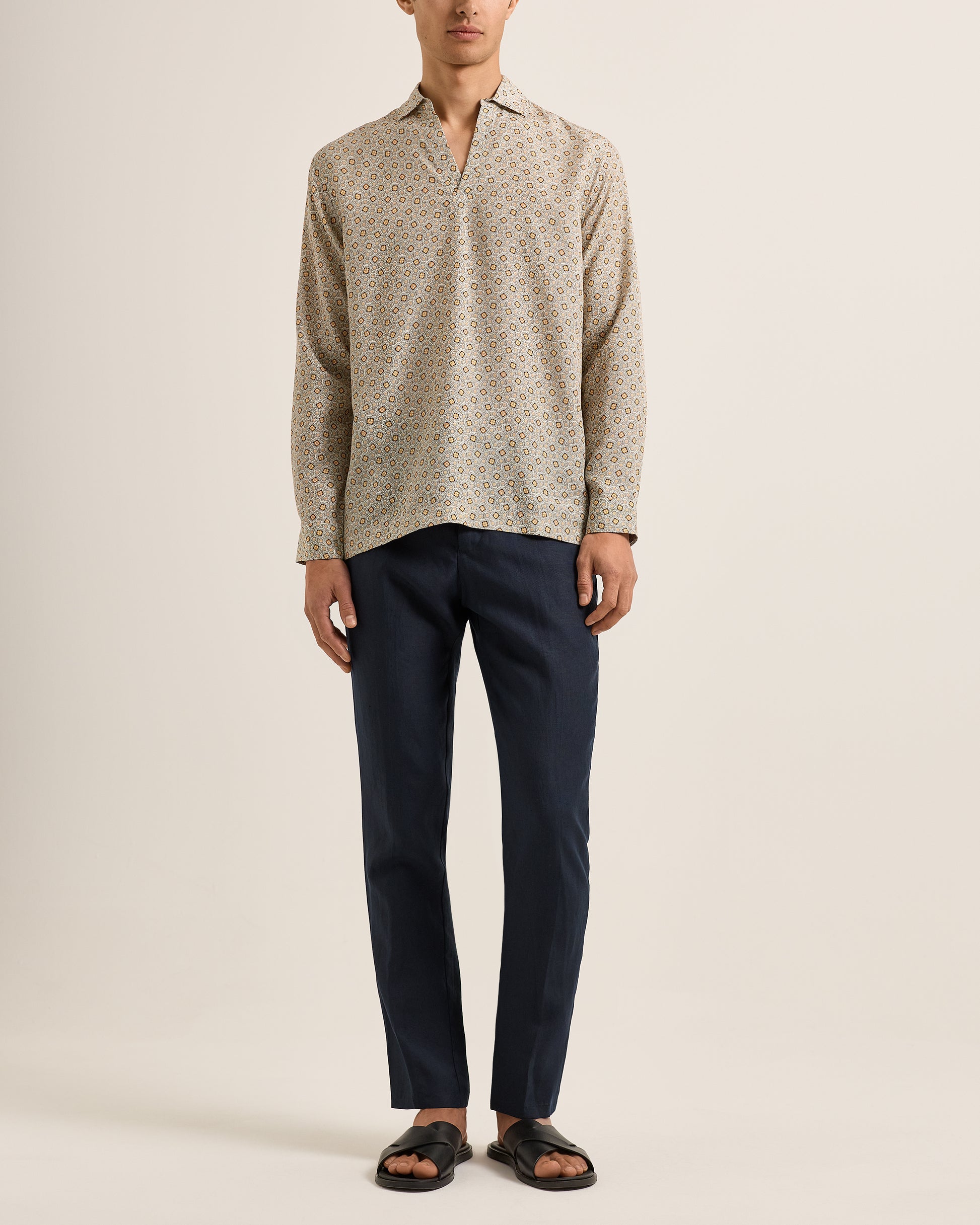 Man wearing a patterned beige shirt and dark pants on a plain background