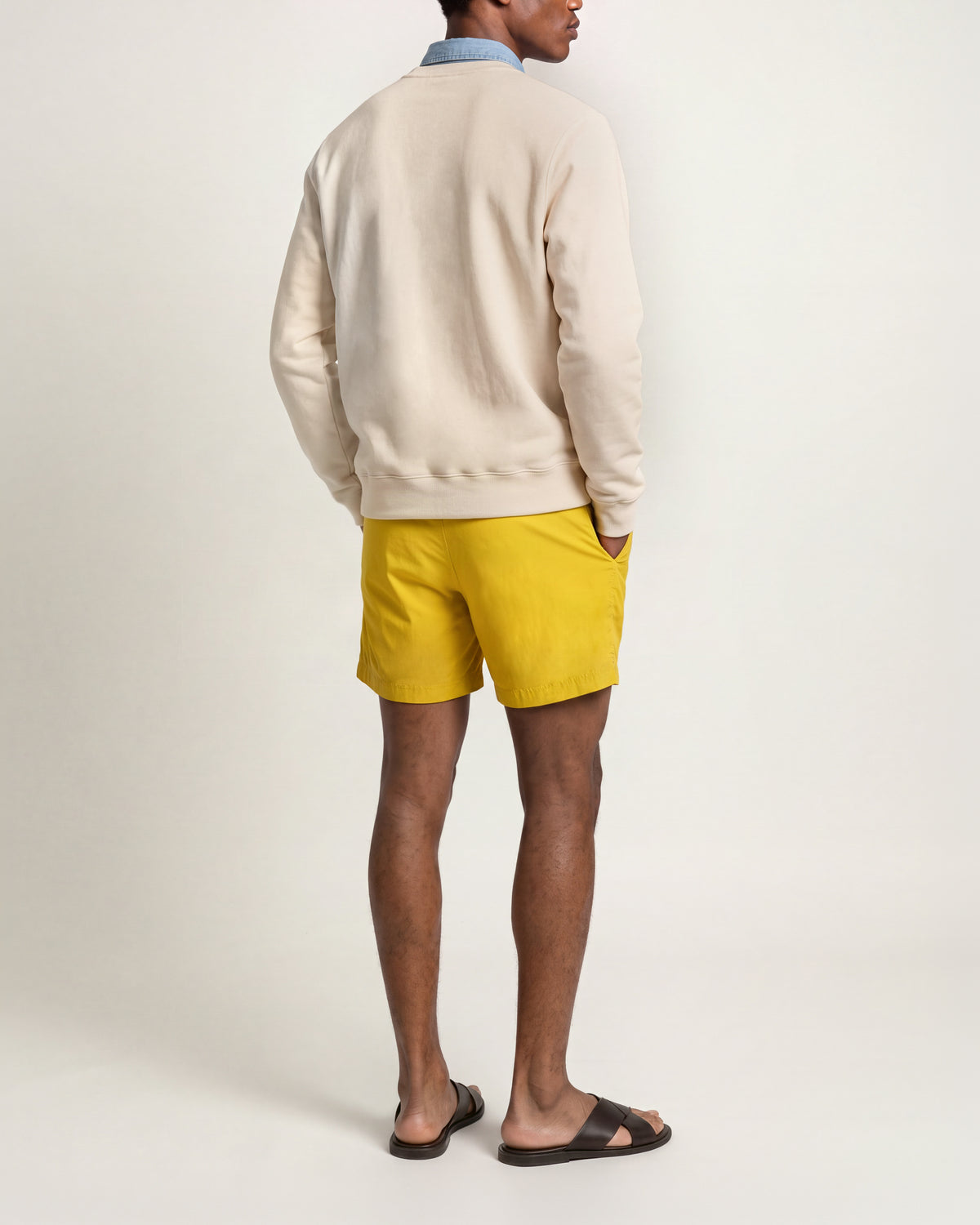 Man wearing light beige sweatshirt with yellow swim shorts and brown sandals on a plain background