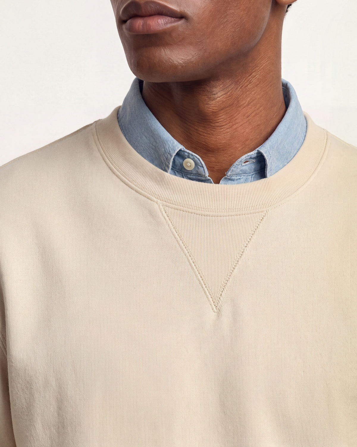 Man wearing beige sweatshirt with denim shirt underneath on a plain background
