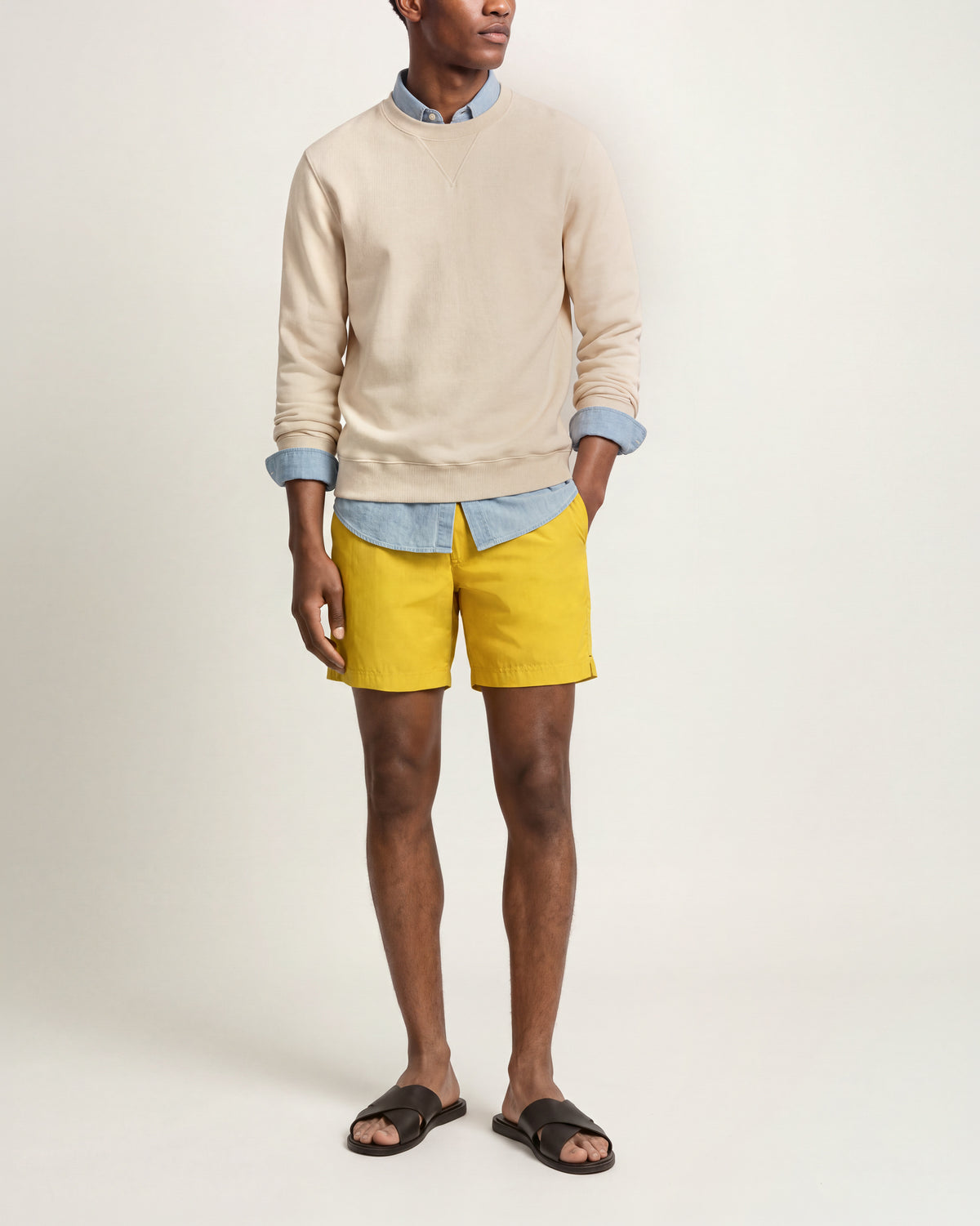 Man wearing beige sweatshirt with denim shirt underneath and yellow swim shorts on a plain background
