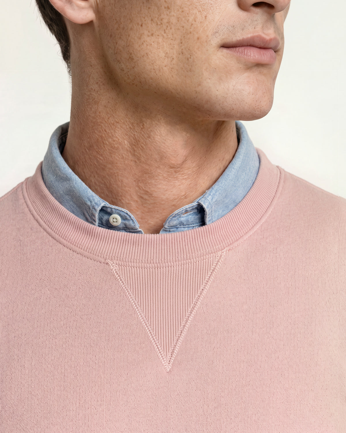Closeup of man wearing light pink sweatshirt with denim shirt underneath on a plain background