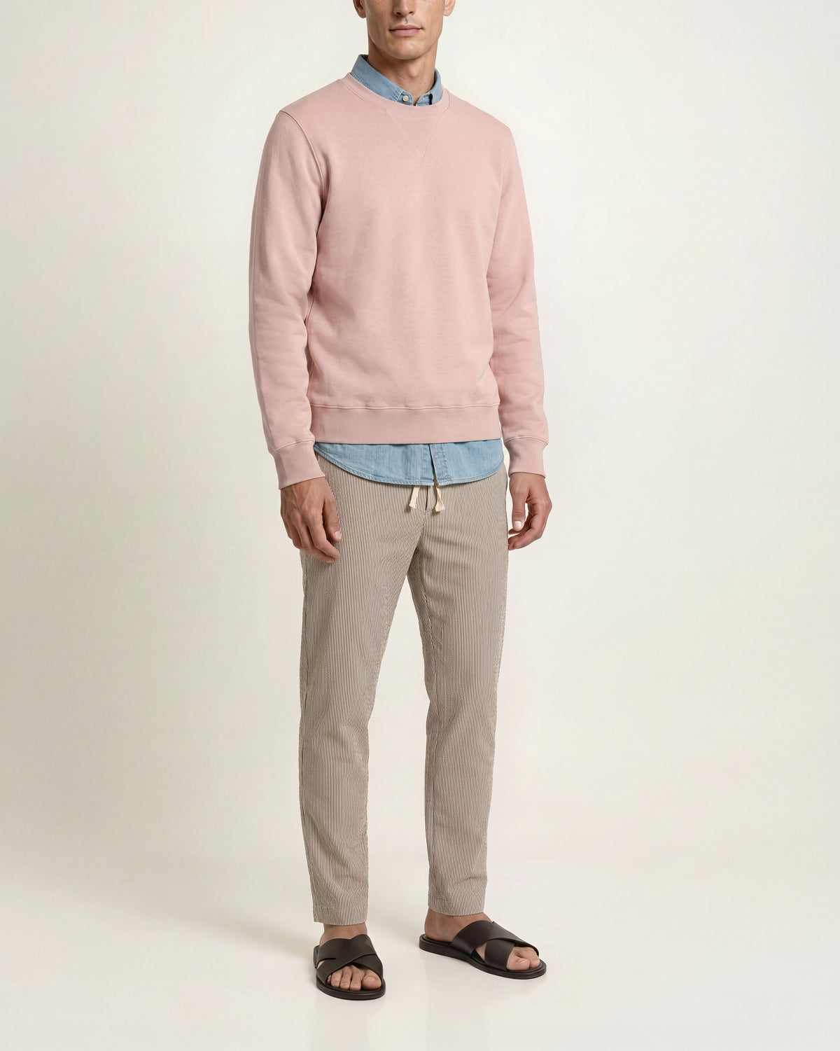 Man wearing pink sweatshirt with denim shirt underneath and brown/ white striped trousers on a plain background