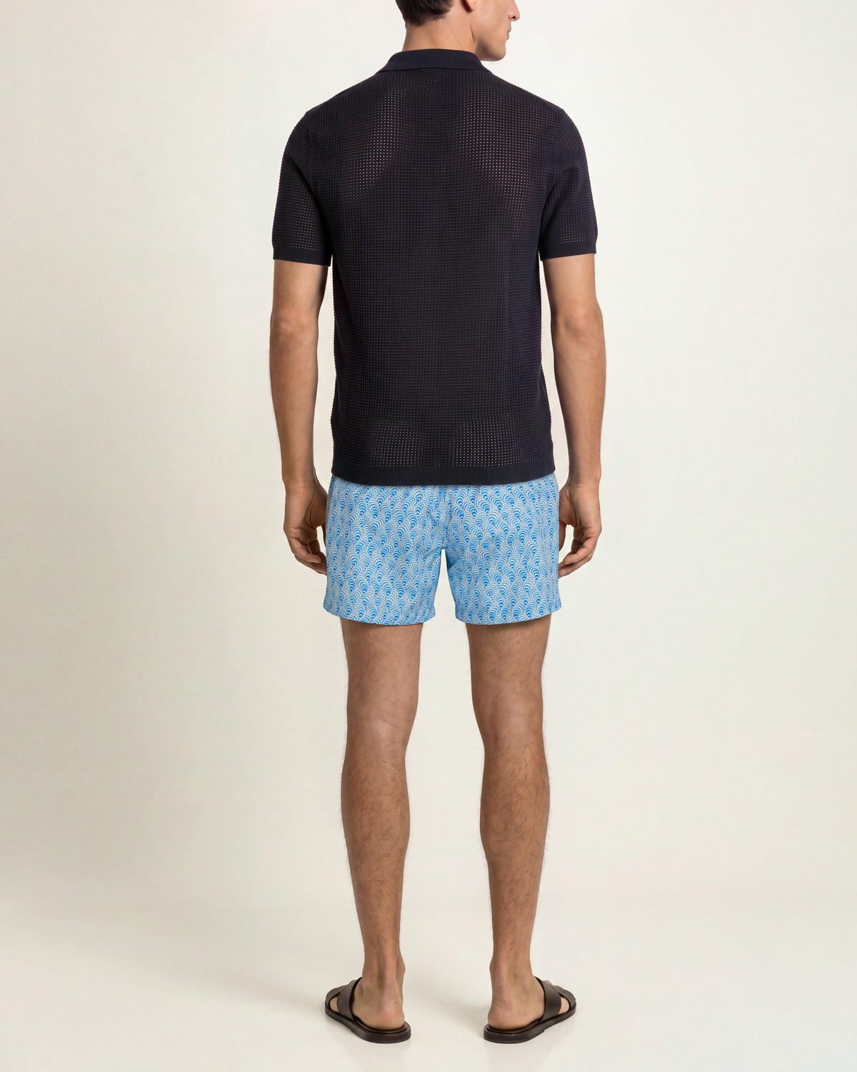 Man wearing navy mesh stitch polo shirt and blue patterned swim shorts on a plain background