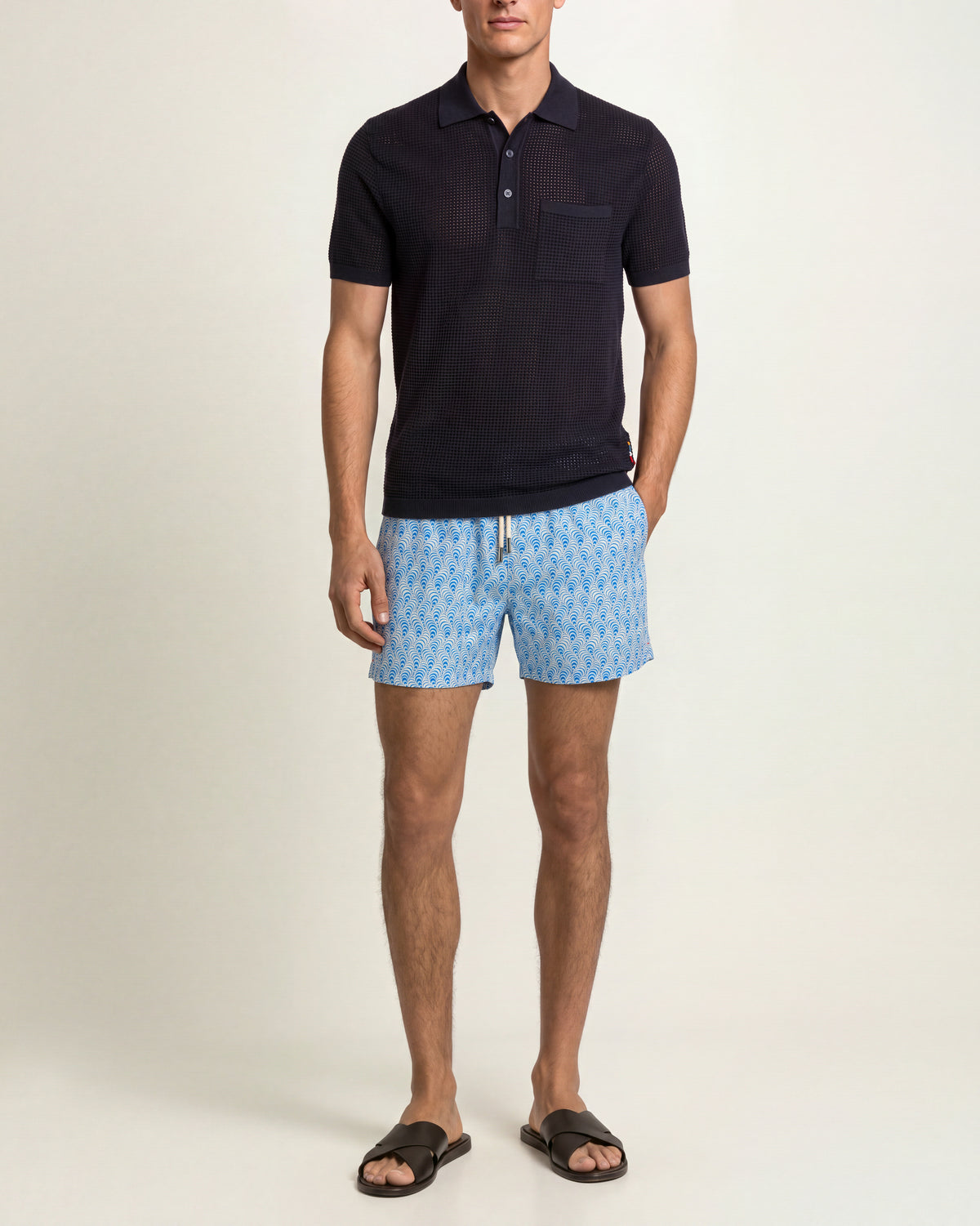 Man wearing navy mesh stitch polo shirt and blue patterned swim shorts on a plain background