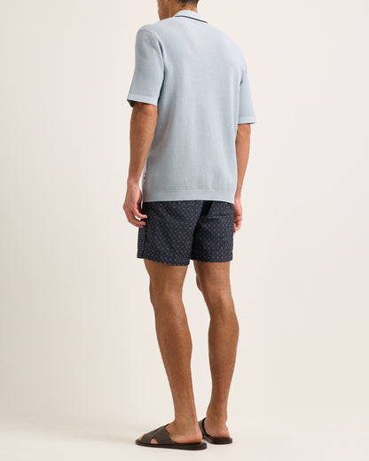 Person wearing a light blue polo shirt and dark patterned shorts on a plain background