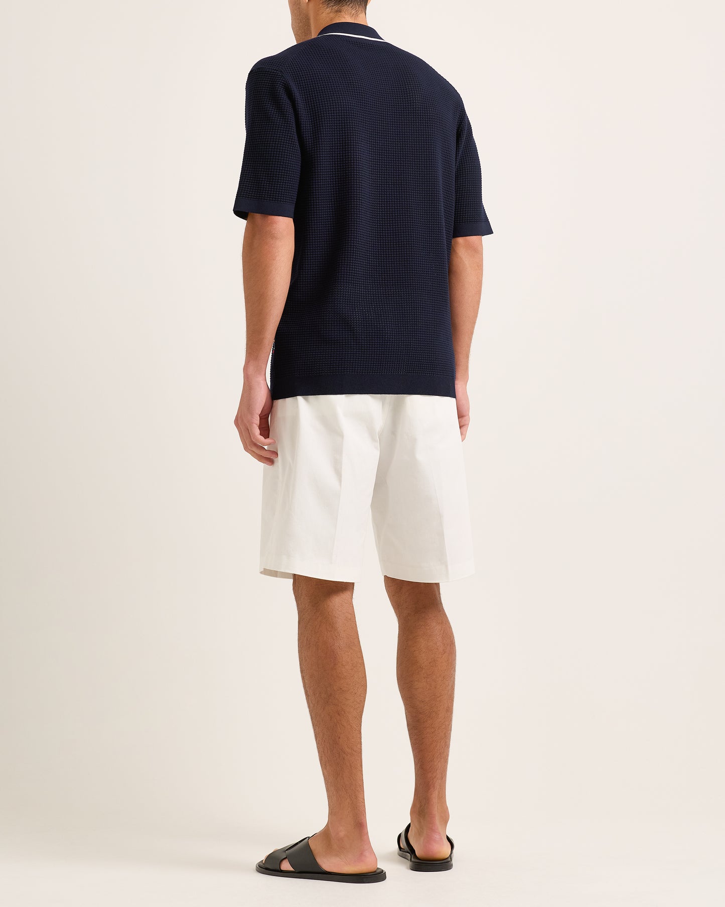 Man wearing navy knitted shirt with white trim and white shorts on a plain background