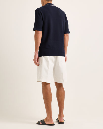 Man wearing navy knitted shirt with white trim and white shorts on a plain background