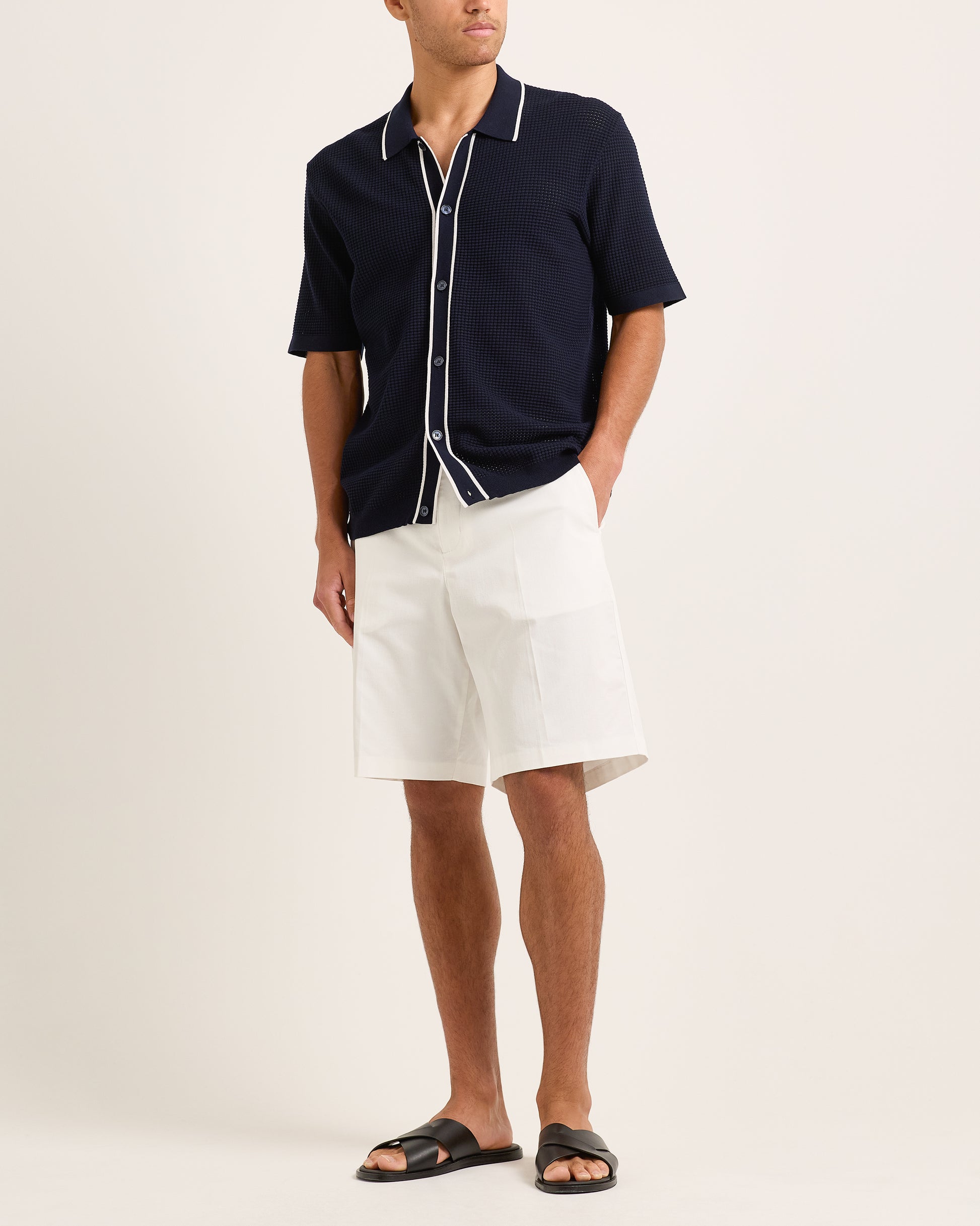Man wearing navy knitted shirt with white trim and white shorts on a plain background