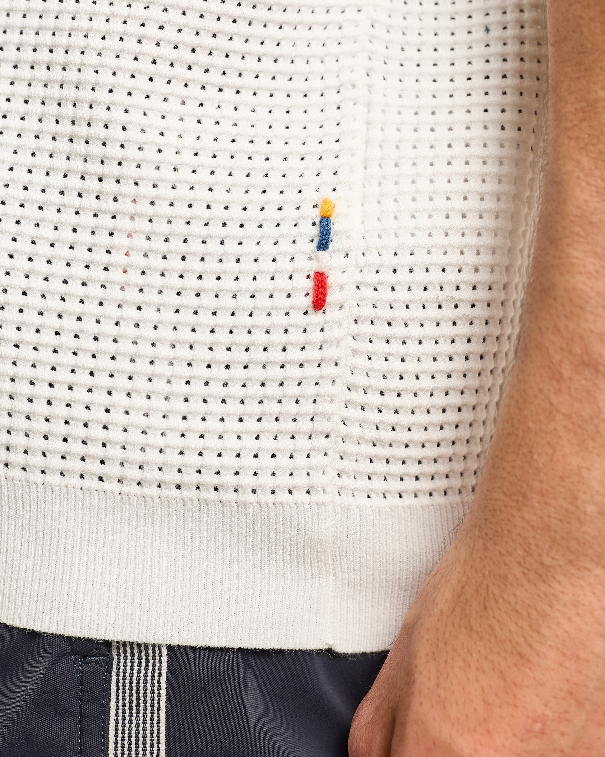 Close-up of a textured white sweater with a small embroidered logo on a person wearing navy pants.