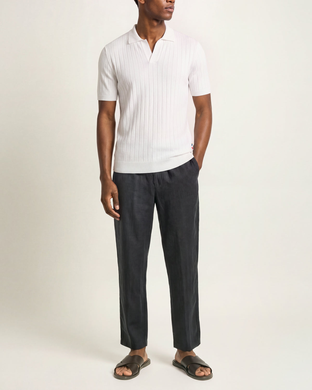 A man modeling a white short-sleeve ribbed knit polo shirt with dark charcoal trousers and dark brown slide sandals, standing against a plain off-white studio background.