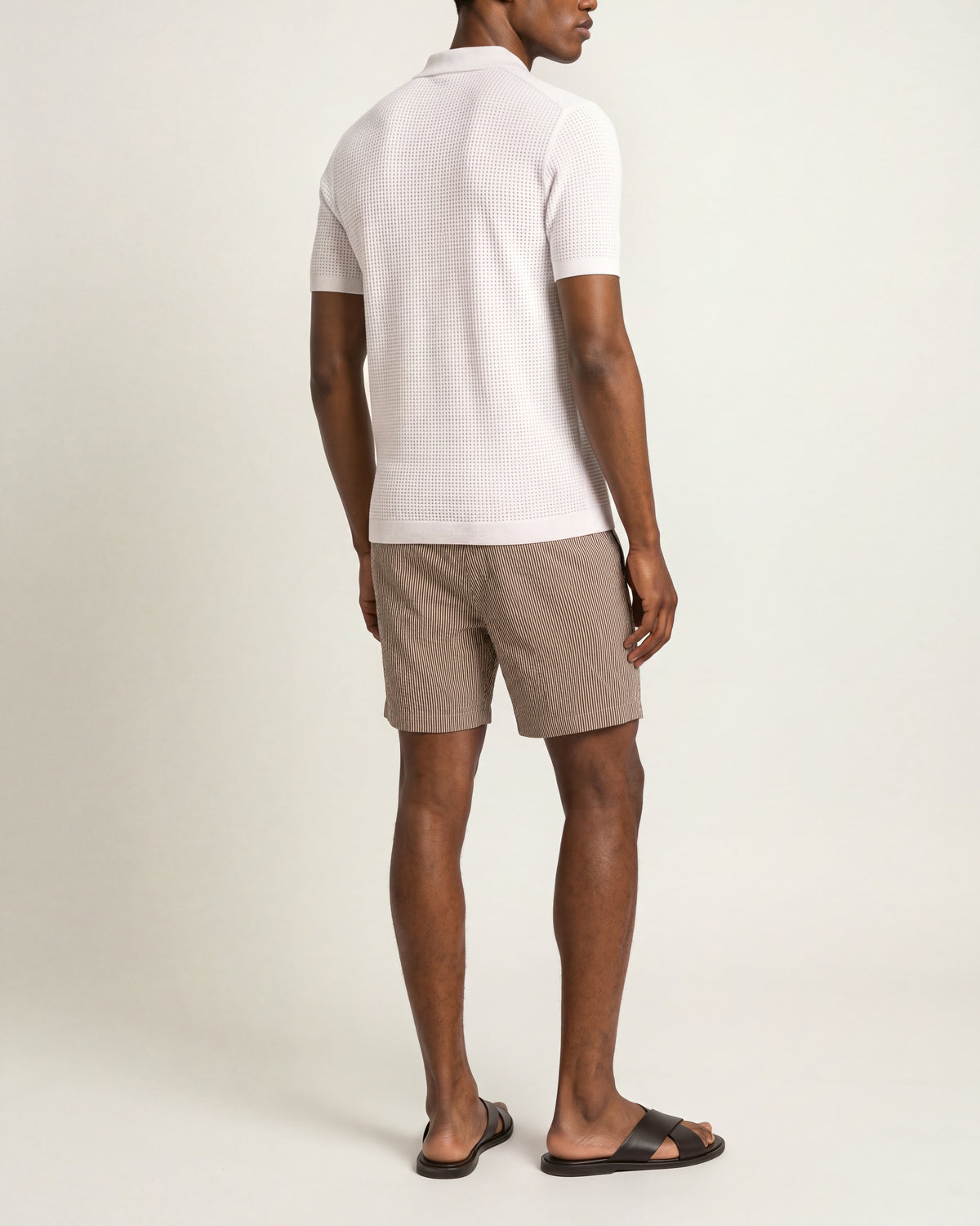 A man shown from the back wearing a white short-sleeve polo shirt, taupe-brown linen shorts, and dark brown slide sandals, photographed against a clean off-white studio background.