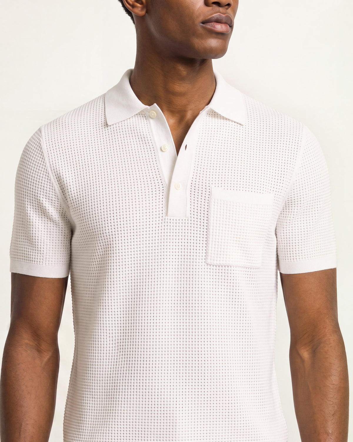 A man wearing a short-sleeve polo shirt with a textured, waffle-like knit pattern in off-white. The photo is a close-up crop of the upper body, showing the collar, button placket, and chest pocket against a plain light background.