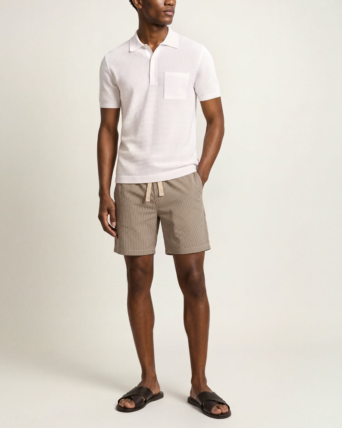 A man modeling a white short-sleeve polo shirt with a chest pocket, paired with beige drawstring shorts and black slide sandals, against a plain light beige studio background.