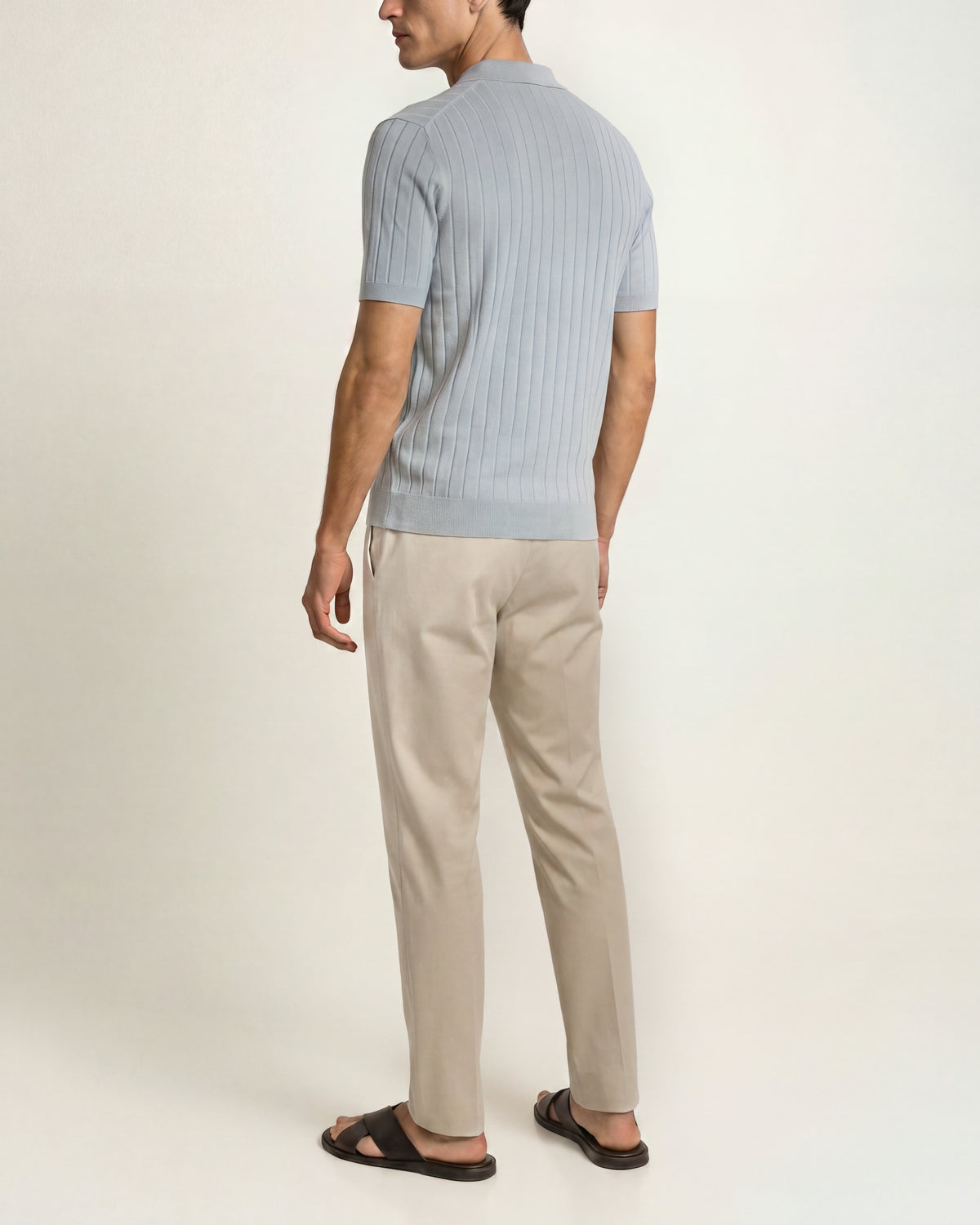 A man shown from the back wearing a light blue short-sleeve ribbed knit polo shirt, beige trousers, and dark brown sandals, standing against a plain off-white studio background.