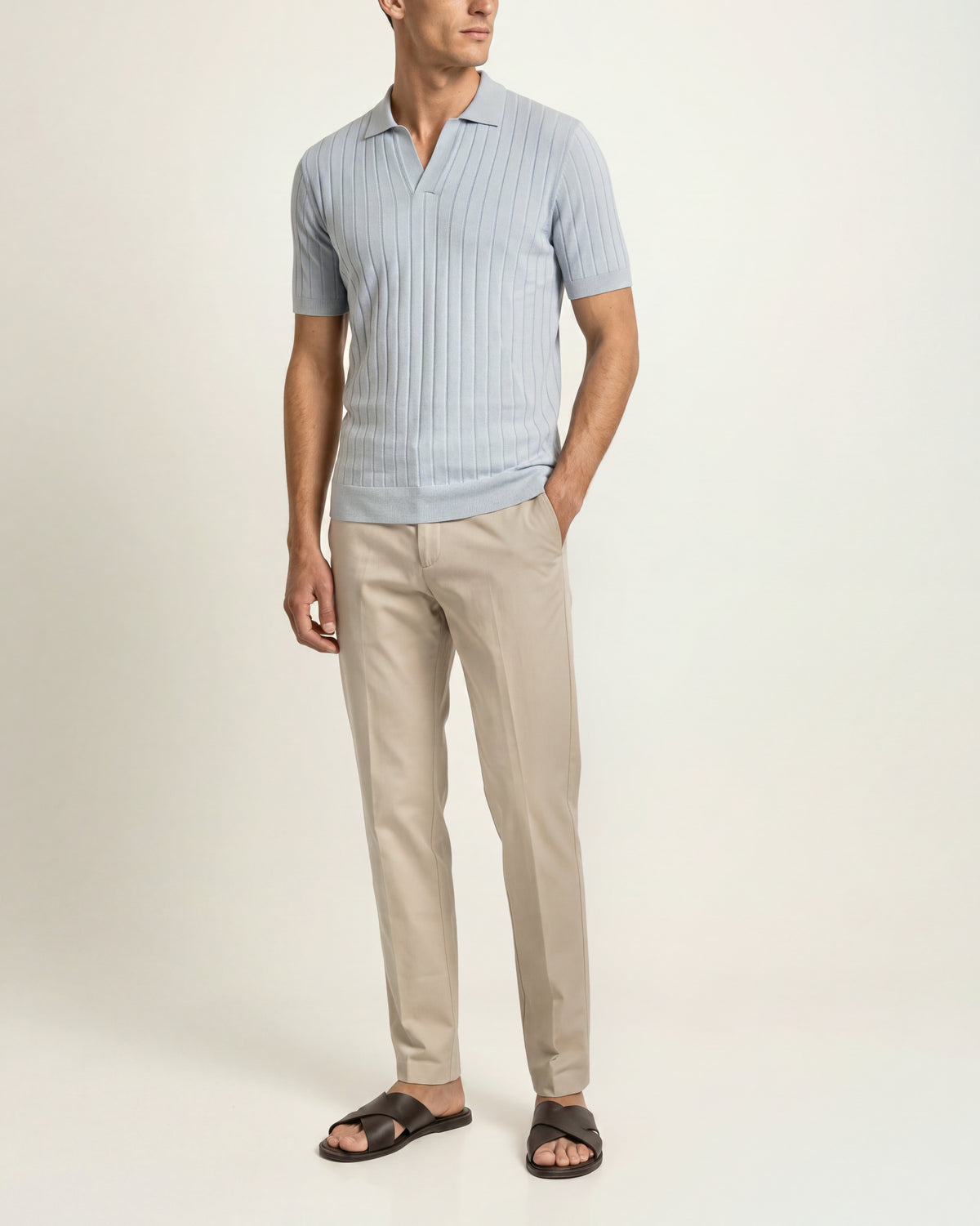 A man modeling a light blue short-sleeve ribbed knit polo shirt paired with beige trousers and dark brown slide sandals, photographed against a clean off-white studio background.