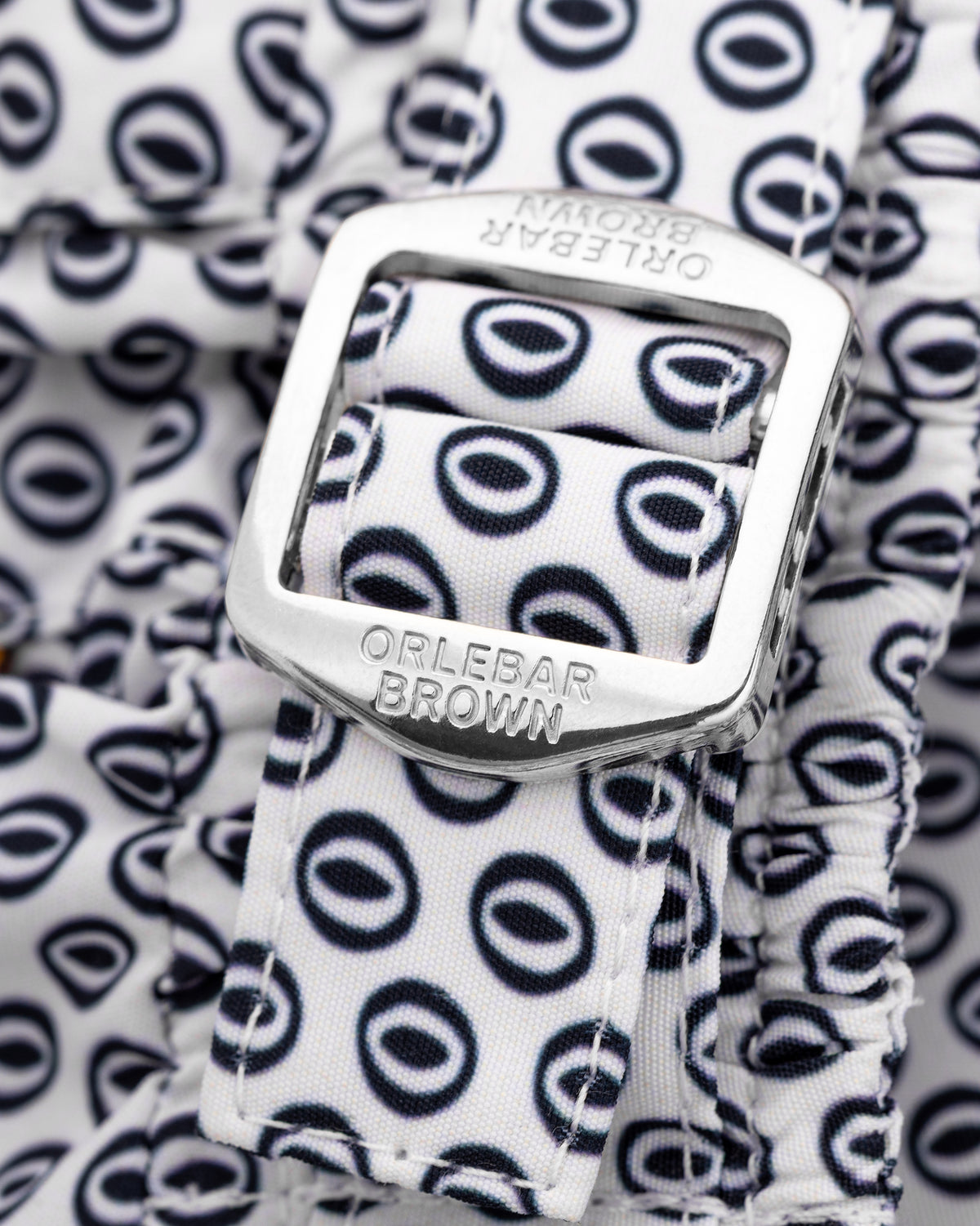 Close-up of a white fabric strap with a repeating navy blue circular logo pattern and a silver metal buckle engraved with the brand name 'ORLEBAR BROWN'.