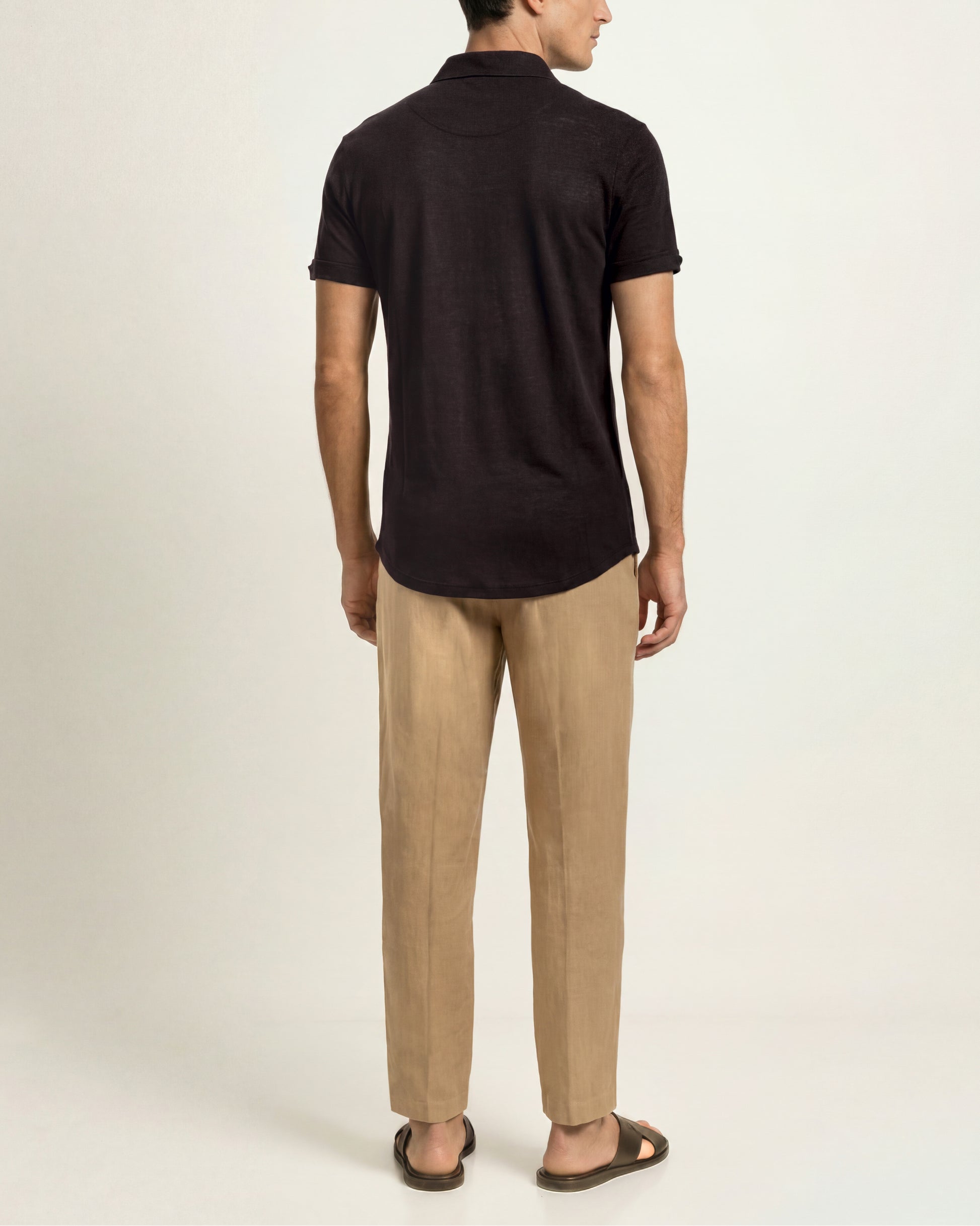 A man shown from the back wearing a dark brown short-sleeve polo shirt, beige trousers, and brown sandals against a plain off-white studio background.