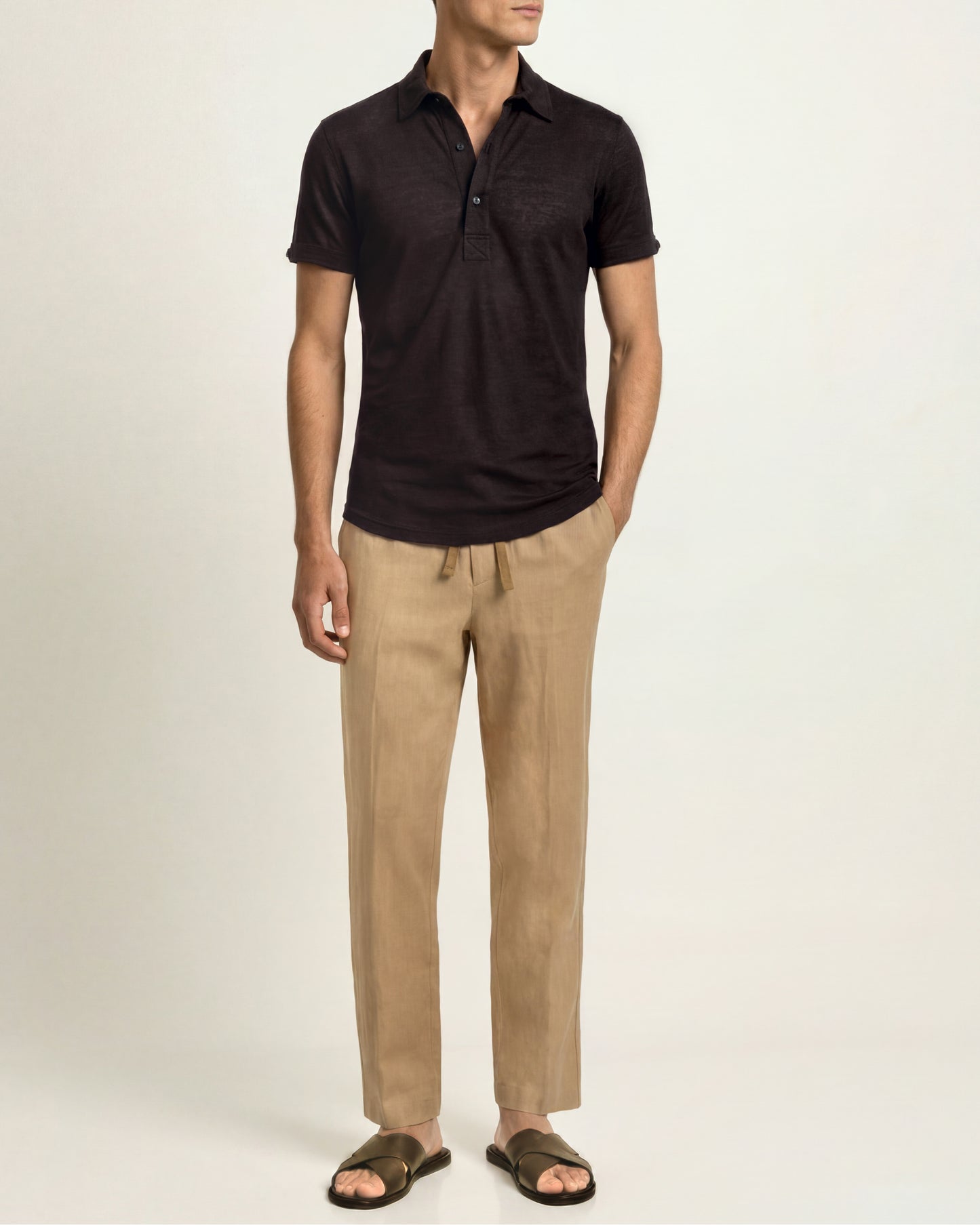 A man wearing a dark brown short-sleeve polo shirt, beige drawstring trousers, and dark brown slide sandals, standing against a plain light beige studio background.