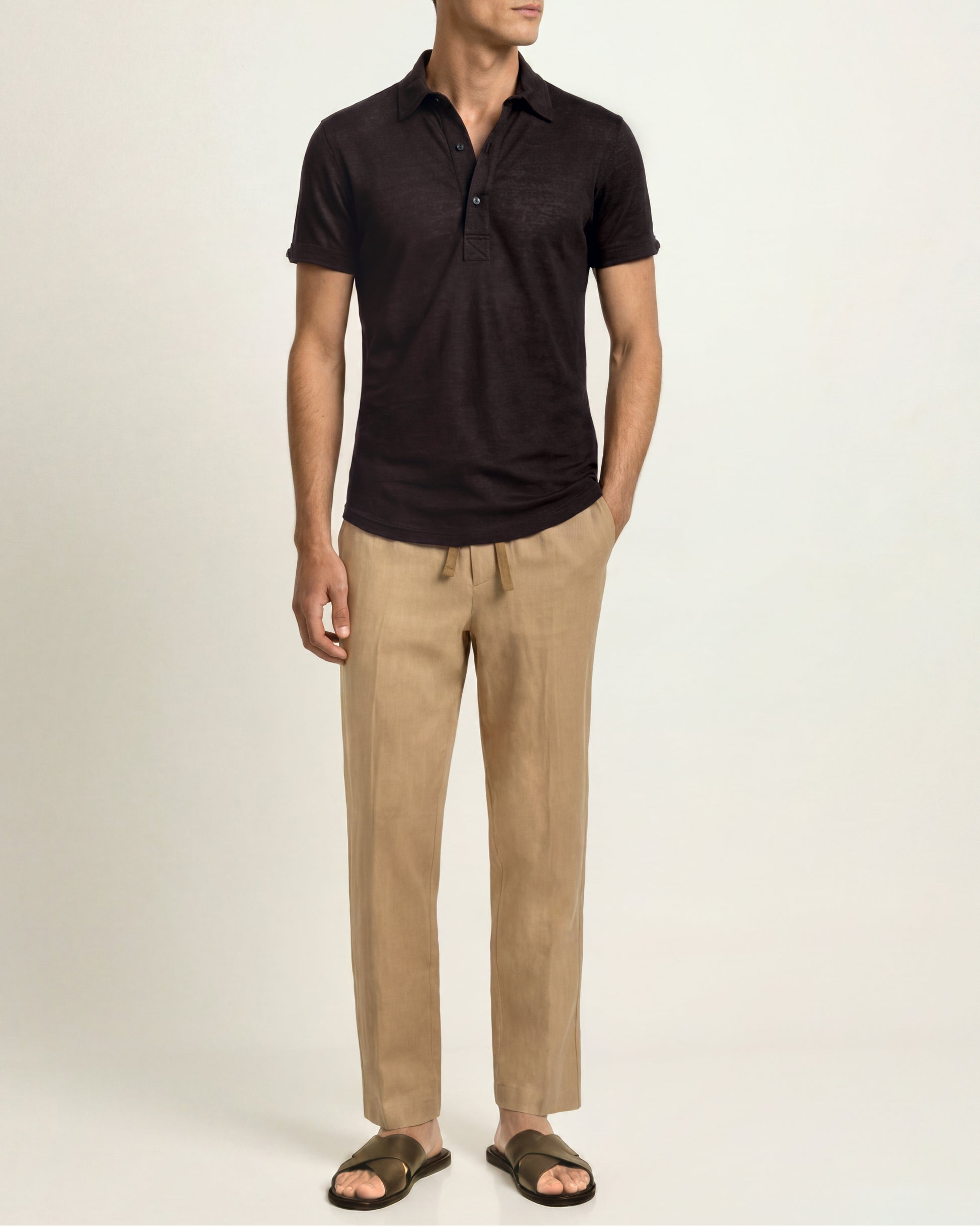 A man wearing a dark brown short-sleeve polo shirt, beige drawstring trousers, and dark brown slide sandals, standing against a plain light beige studio background.