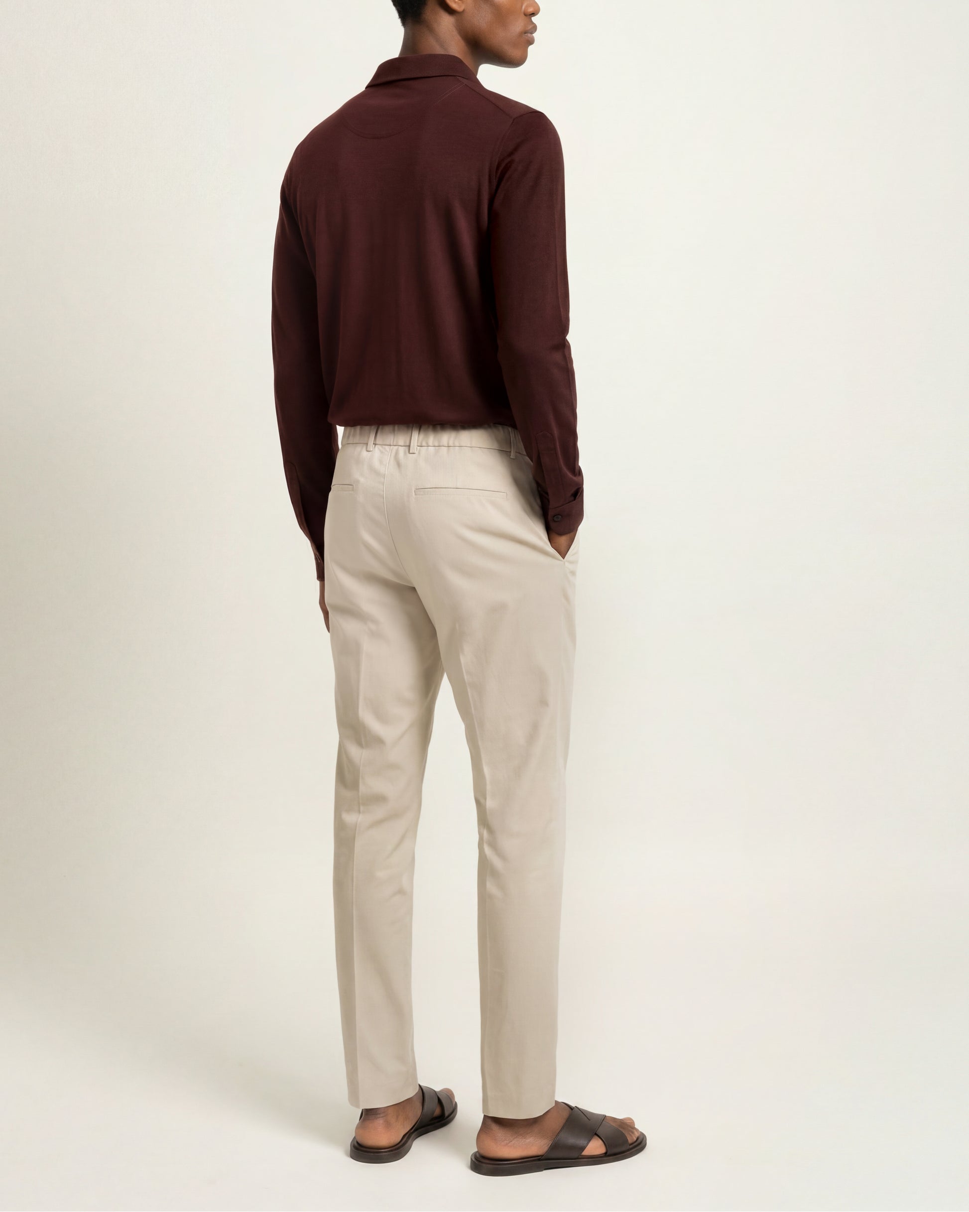 A man shown from the back wearing a dark burgundy long-sleeve polo shirt, light beige trousers, and dark brown sandals against a plain off-white studio background.