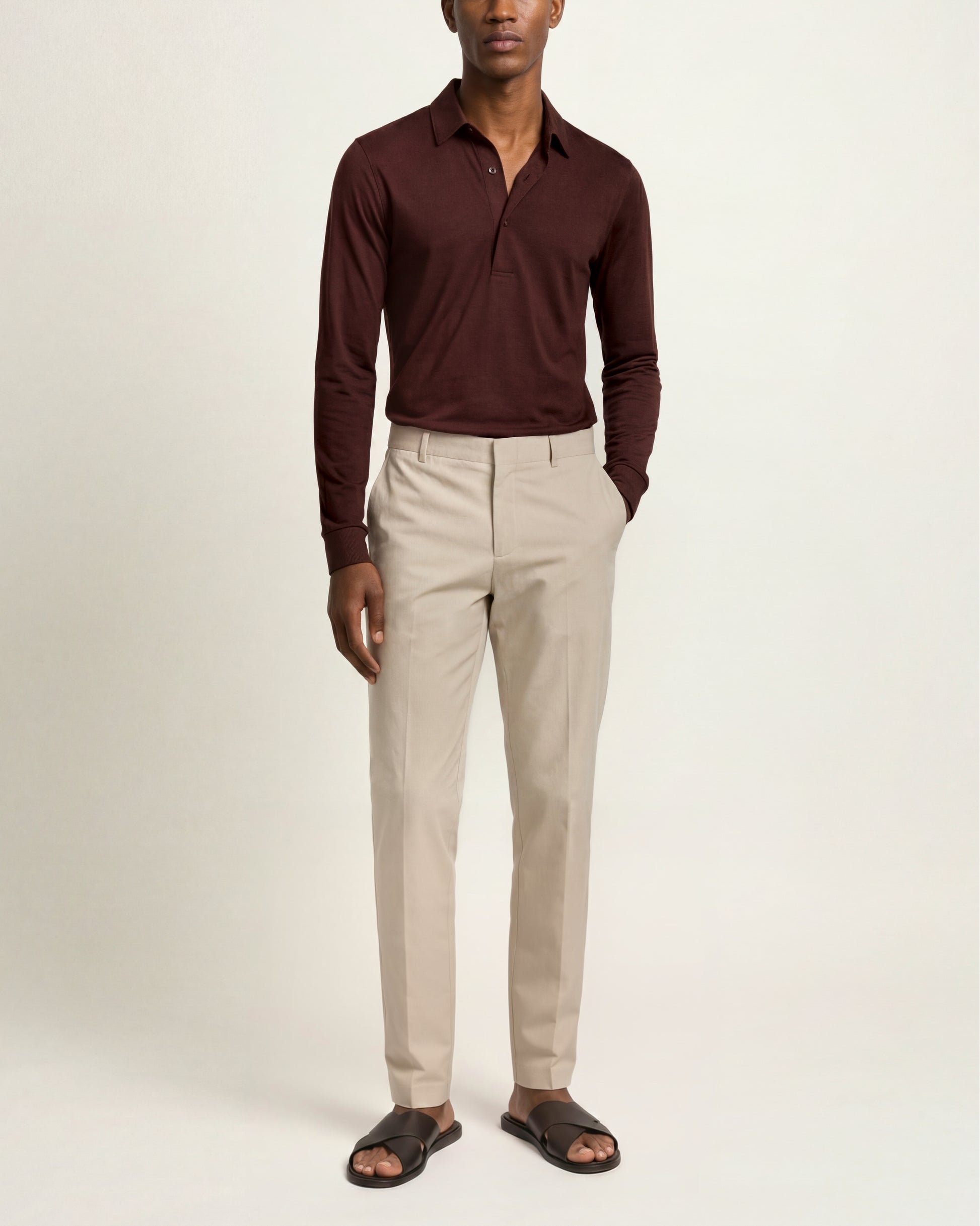A man modeling a dark burgundy long-sleeve polo shirt tucked into light beige trousers, paired with dark brown slide sandals, against a plain off-white studio background.