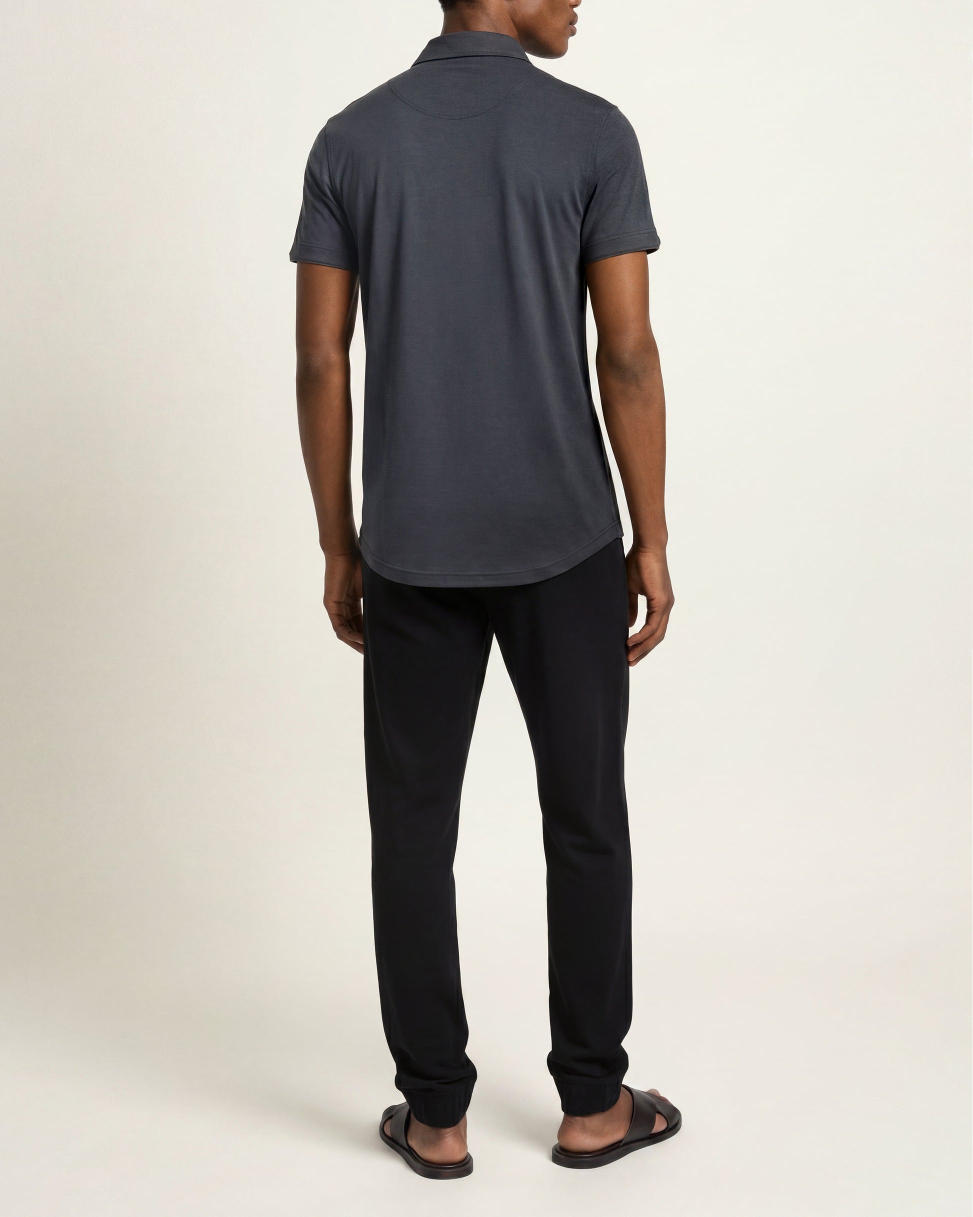 A man shown from the back wearing a dark charcoal gray short-sleeve polo shirt, black trousers, and black sandals, standing against a plain off-white studio background.