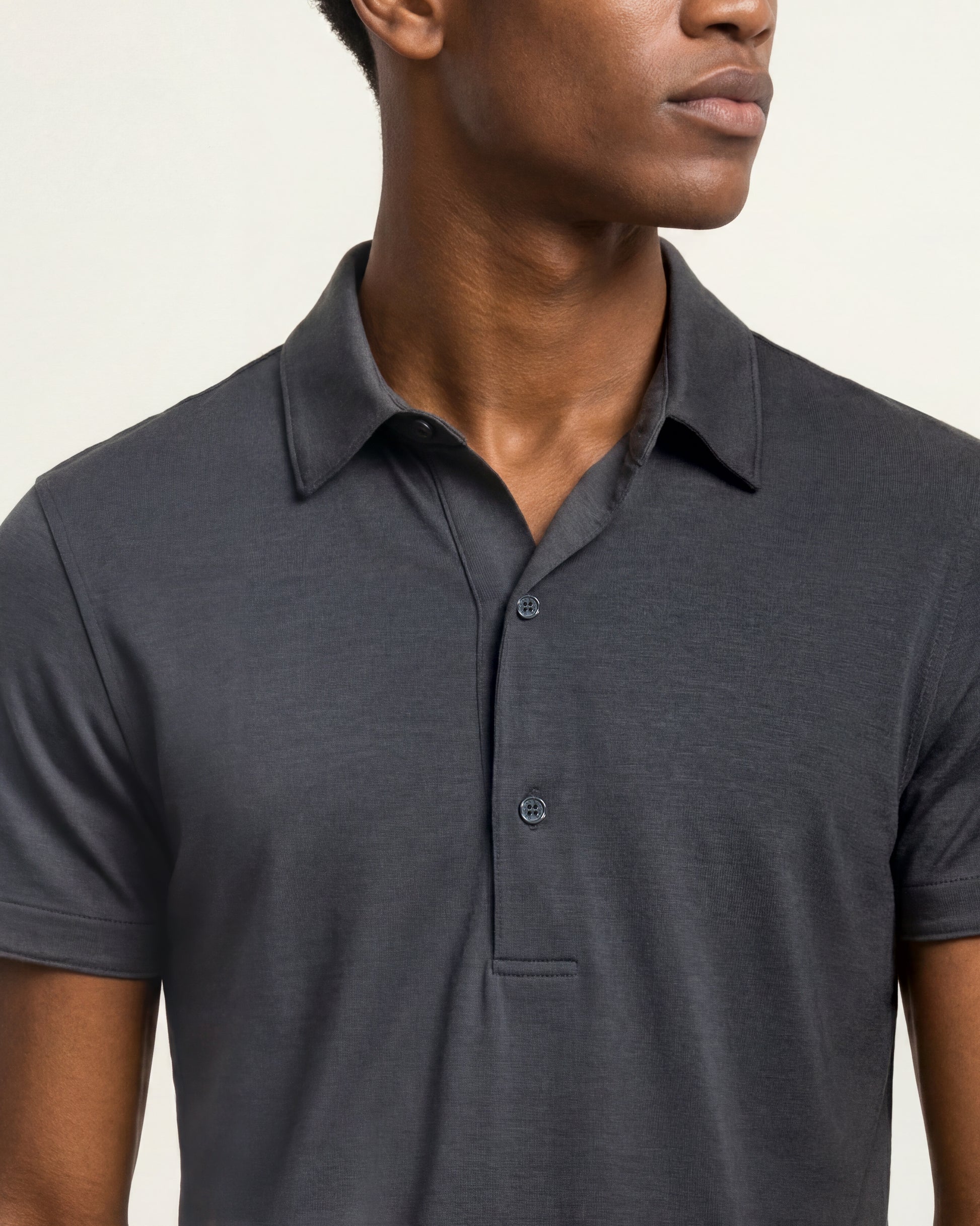 A close-up of a man wearing a dark charcoal gray short-sleeve polo shirt against a light beige background.