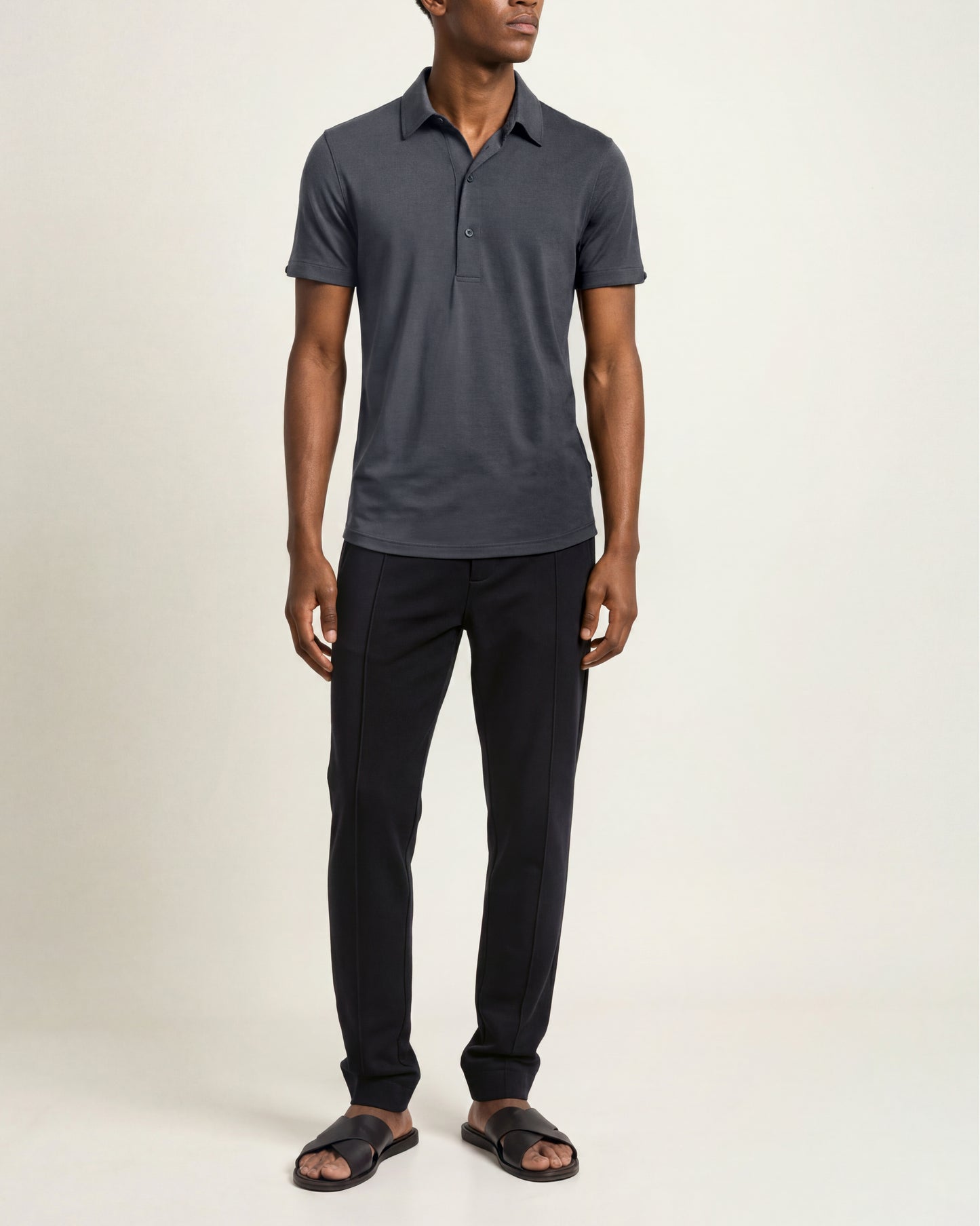 A man modeling a dark gray short-sleeve polo shirt with black trousers and dark brown slide sandals, standing against a plain off-white studio background.
