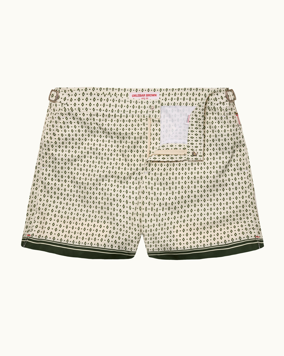A pair of men's shorts featuring an all-over small black monogram pattern on a cream-colored fabric, with a dark green striped inner hem and a white paper brand tag attached to the back pocket.