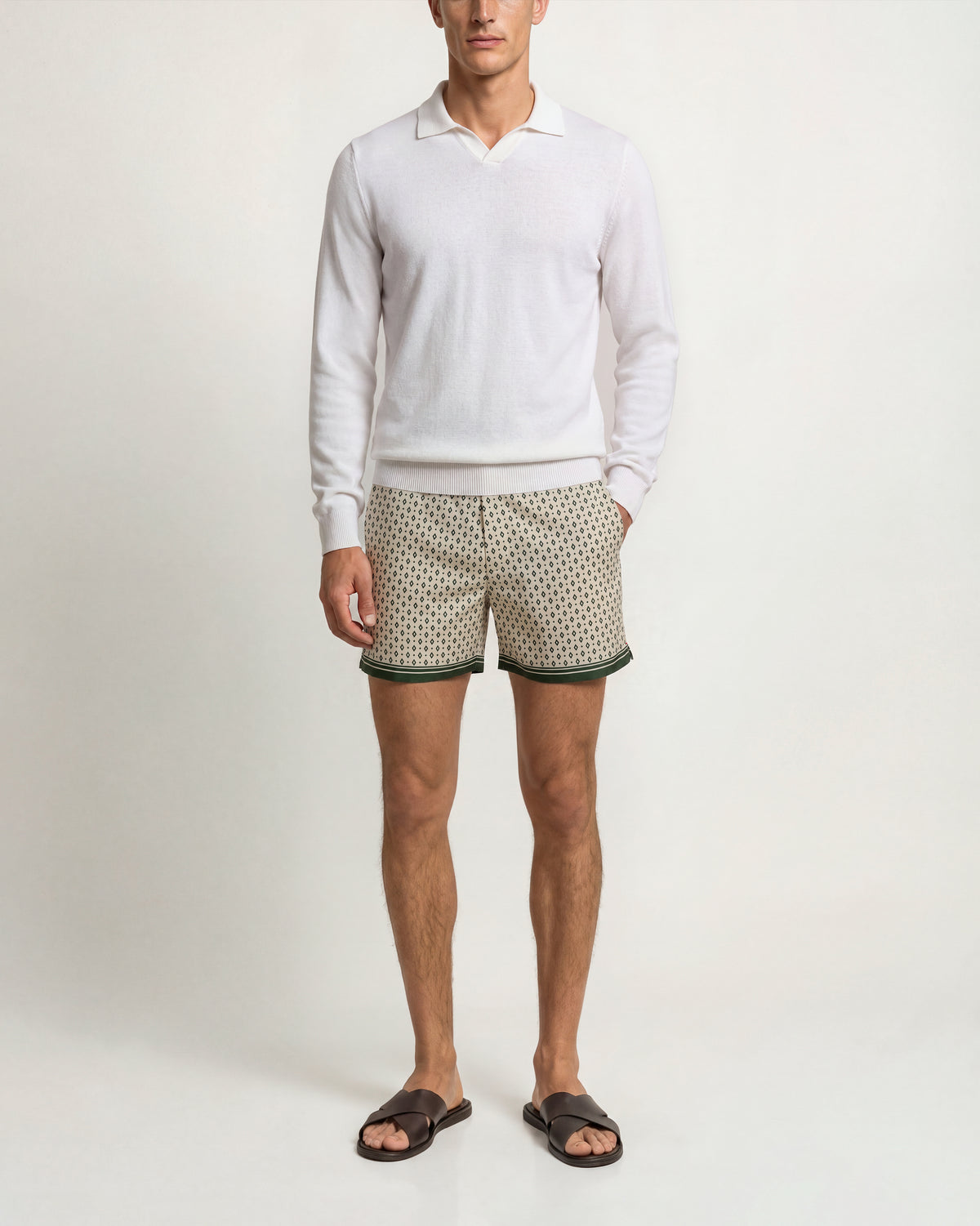 A man modeling a white long-sleeve polo shirt and beige shorts with a green and white geometric pattern and green trim, paired with dark brown slide sandals, against a plain off-white studio background.