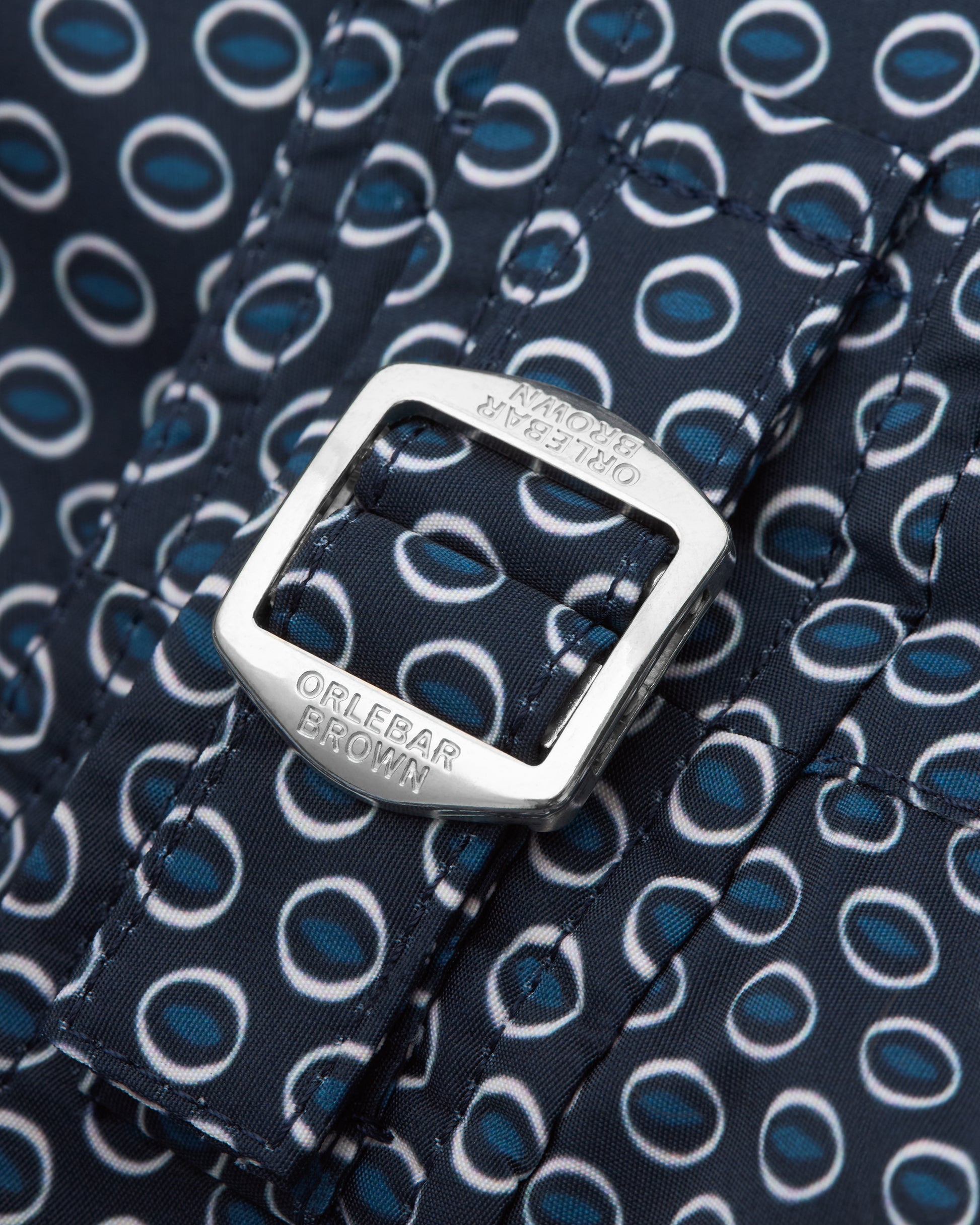 Close-up of a belt with a navy blue and white circular pattern, featuring a silver-tone metal buckle engraved