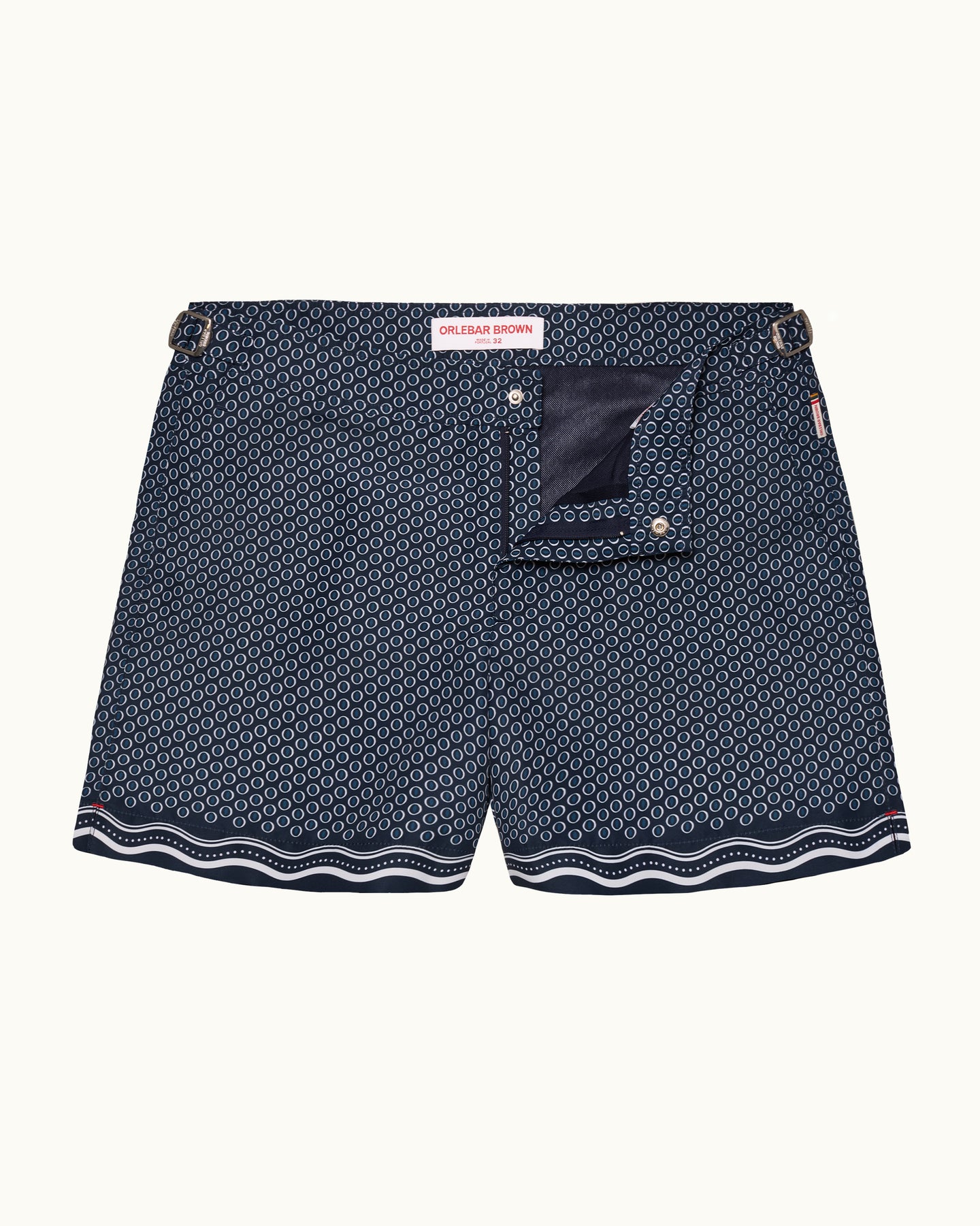 A pair of men's swimshorts featuring a navy blue and white geometric pattern, a solid navy back pocket, and a wavy white stripe along the hem, displayed against a light cream background.